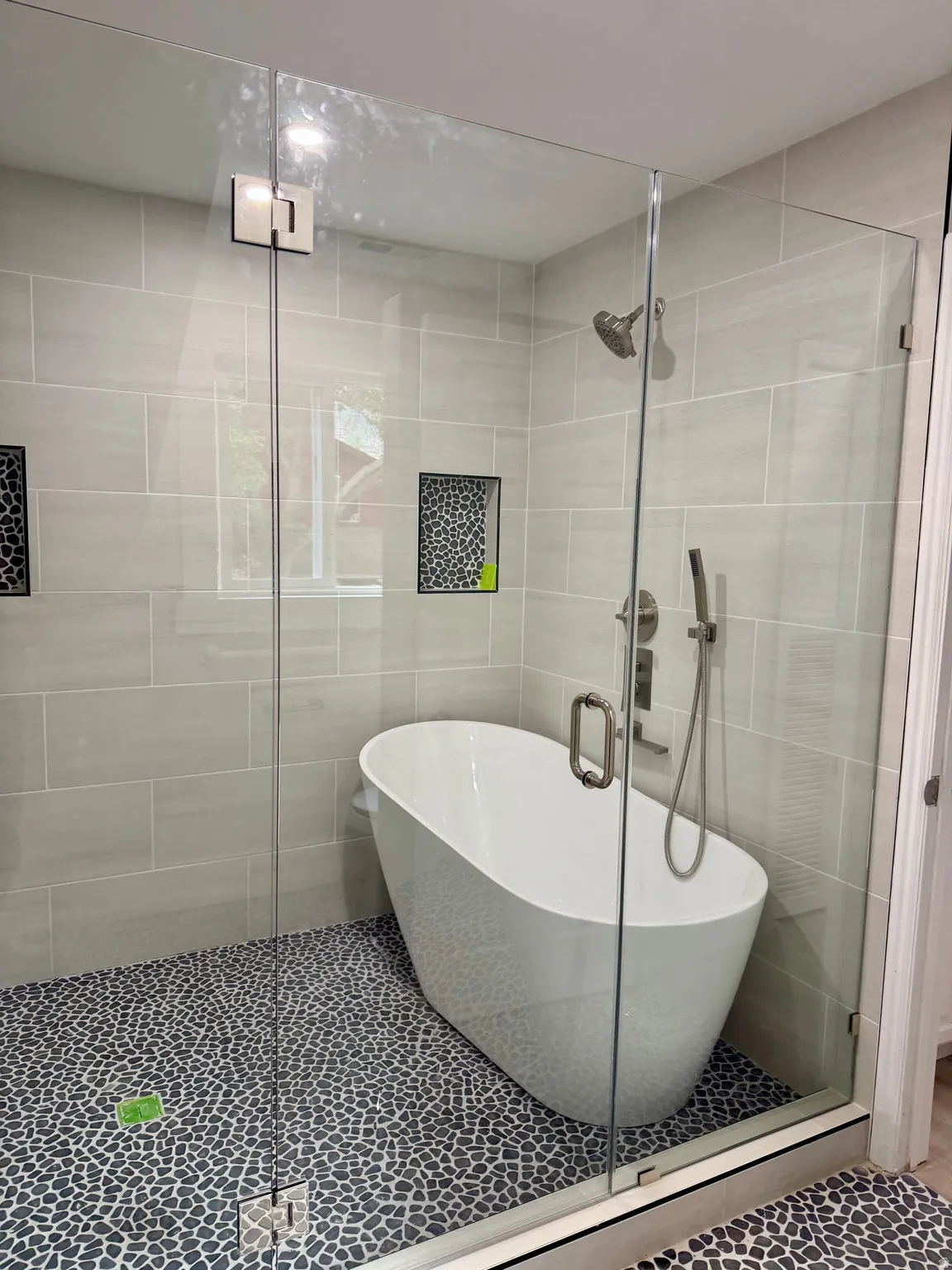 Glass-enclosed wet room featuring a freestanding soaking tub, pebble tile flooring, and light grey subway tile walls