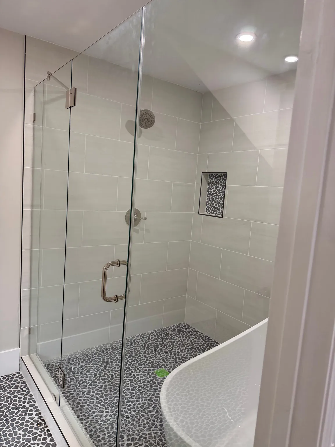 Walk-in shower with clear glass enclosure, light grey subway tile walls, a built-in shower niche with mosaic tile, and a pebble-finish floor