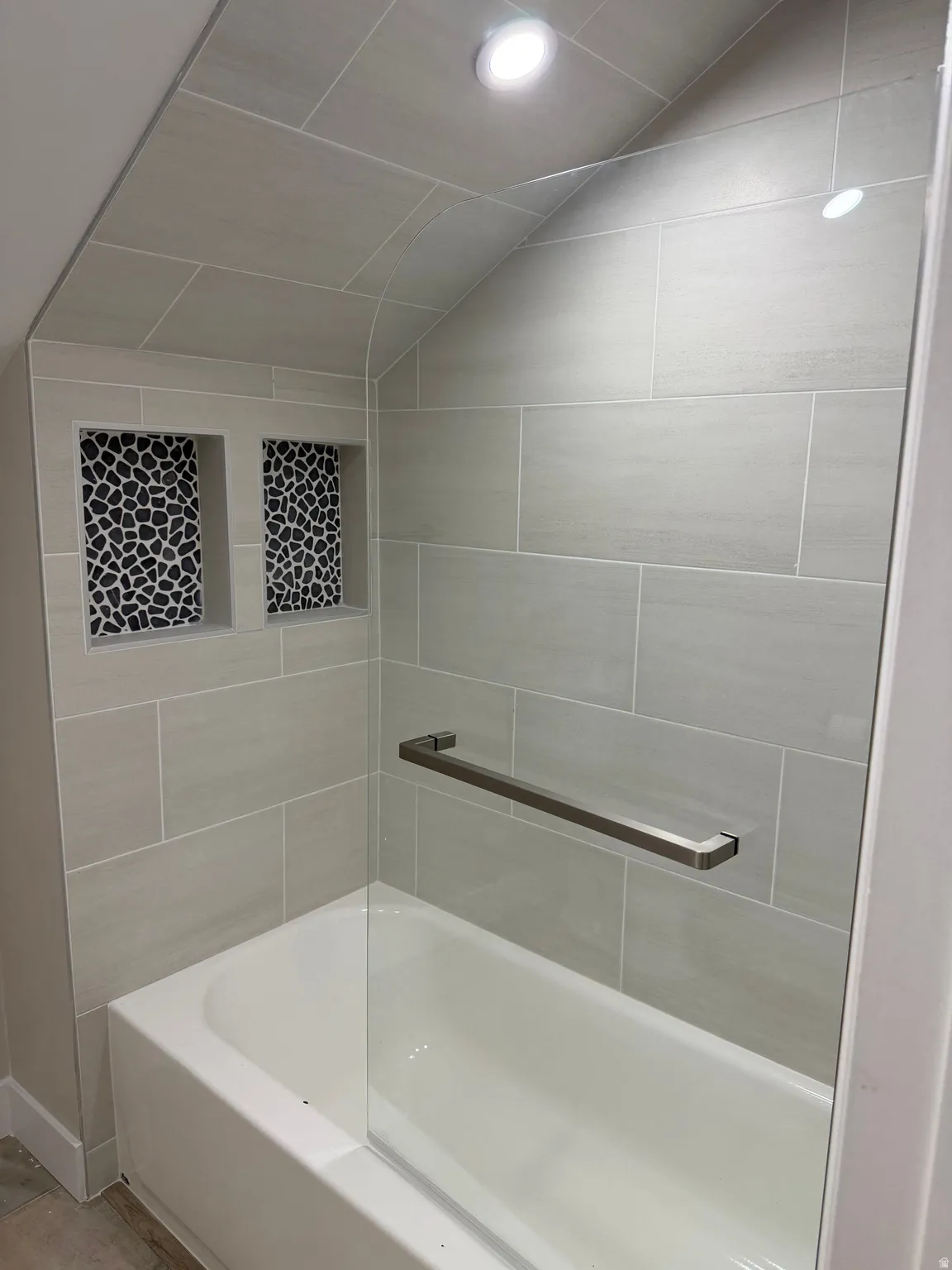Bathtub with light gray subway tile surround, two tiled shower niches with black and white pebble mosaic, clear glass shower panel, and recessed lighting