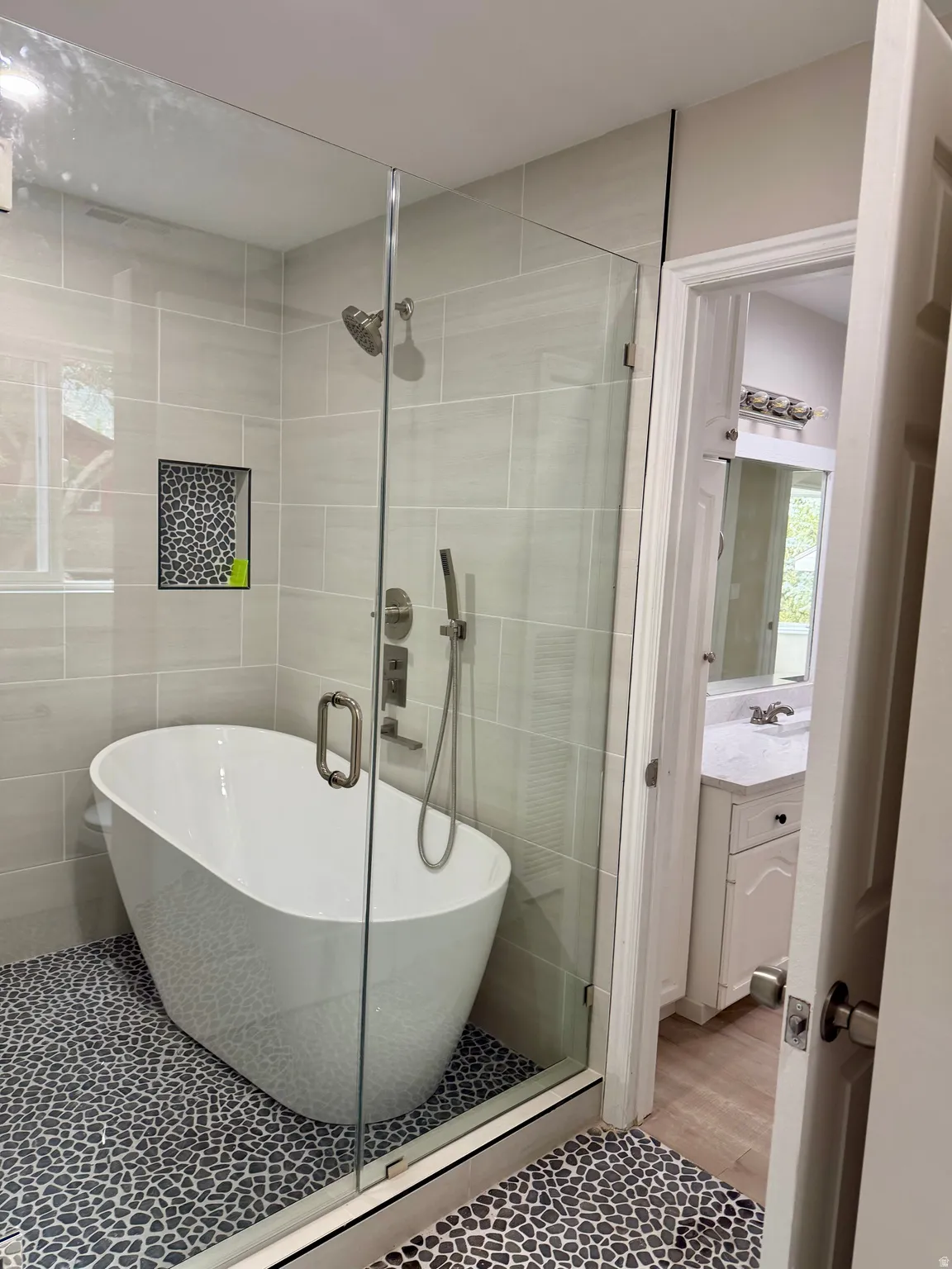 Enclosed shower featuring a freestanding soaking tub, pebble tile flooring, glass shower enclosure, a built-in shower niche, and a fixed showerhead with a handheld sprayer