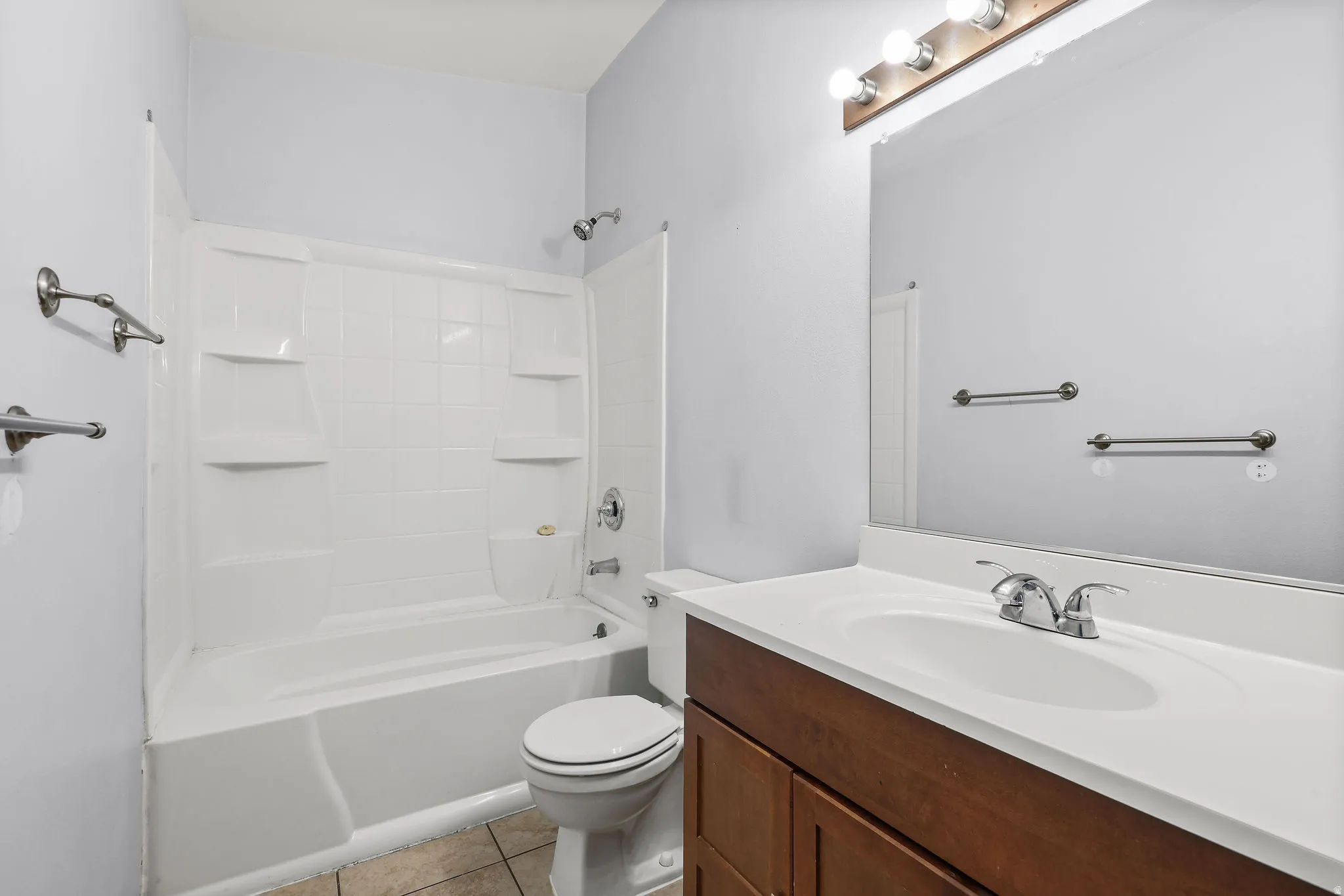 Bathroom featuring a white tub and shower combination with integrated shelving, a wood-finish vanity with a white countertop and integrated sink, a large wall-mounted mirror, and tile flooring