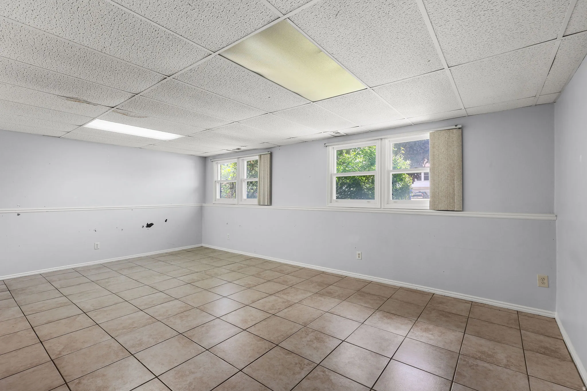 Spacious room featuring neutral tile flooring, light blue walls with white trim, two windows, and a dropped ceiling with recessed lighting