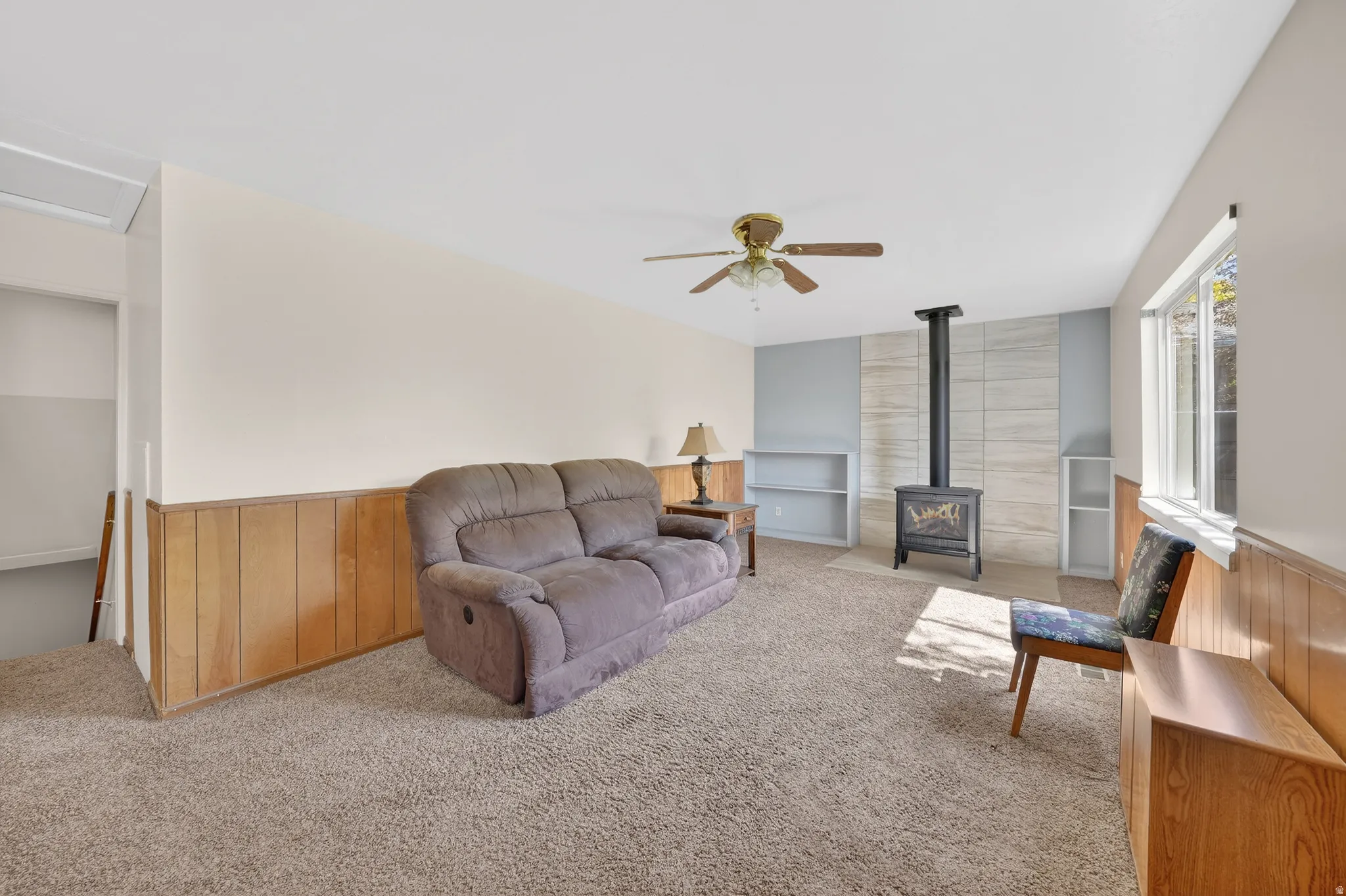 Living room with wooden walls, carpet flooring, a wainscoted wall, a wood stove, and a ceiling fan