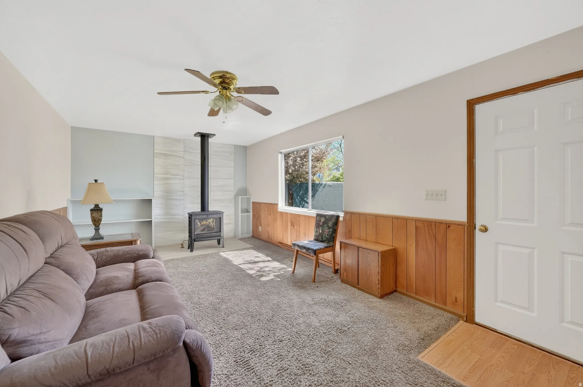 Living room with wood walls, a wood stove, a wainscoted wall, light colored carpet, and a ceiling fan