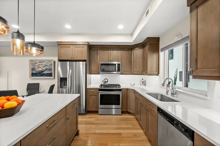 Kitchen with stainless steel appliances, light stone counters, wood finish cabinetry, light wood-style floors, and hanging light fixtures
