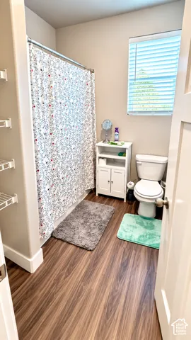 Wood-finish flooring with a white toilet, built-in shelving, and a window with blinds