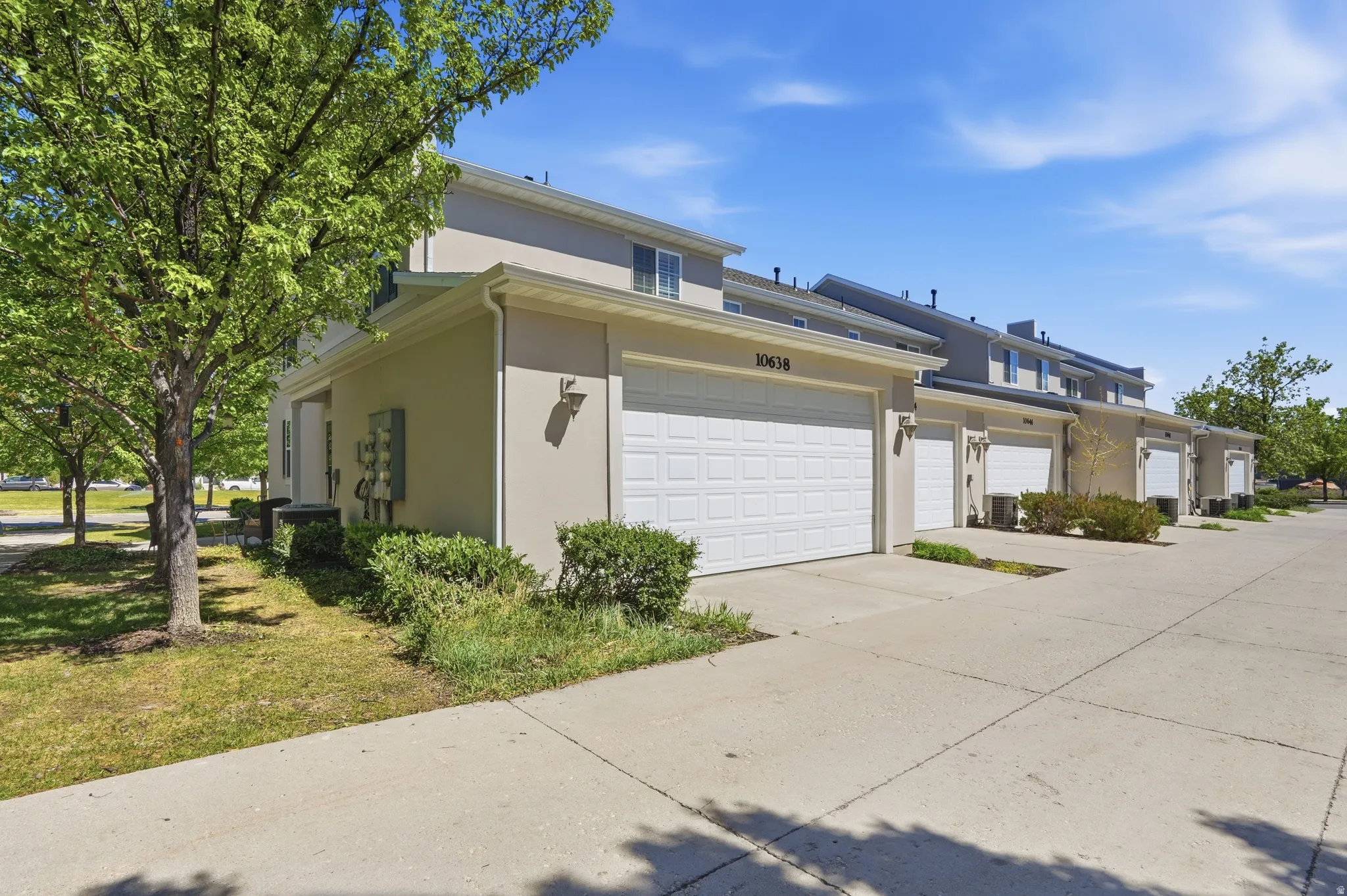 Cream-toned stucco exterior featuring a white garage door, concrete driveway, and mature landscaping