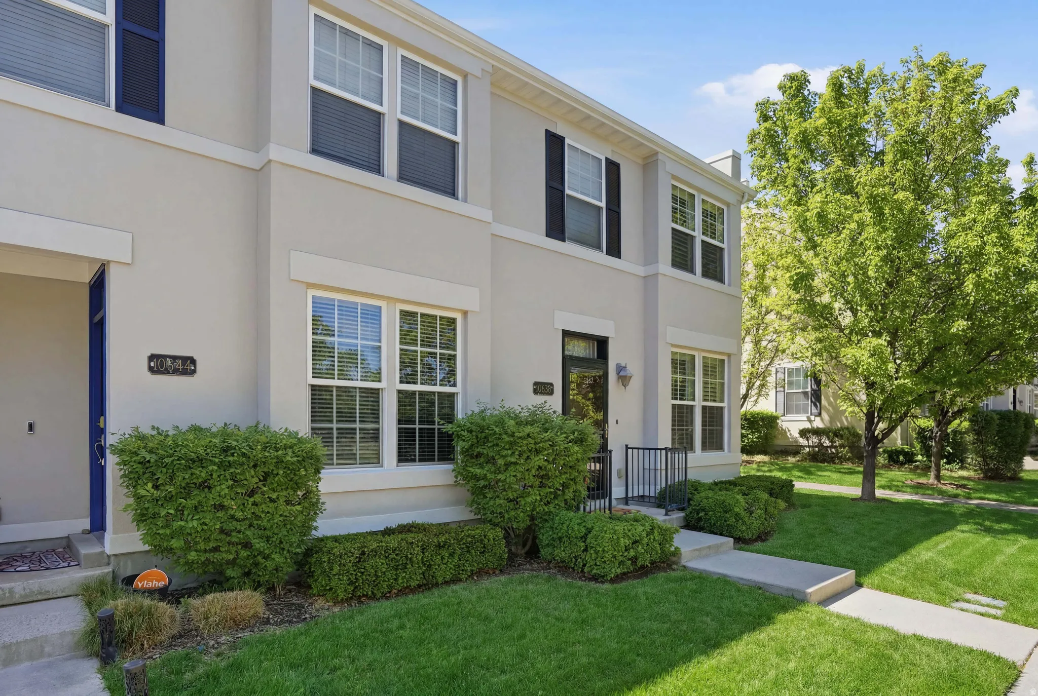 Two-story townhouse featuring a light-colored stucco exterior, multiple windows with white frames, black exterior shutters, a black front door with a glass panel, and a small black metal railing