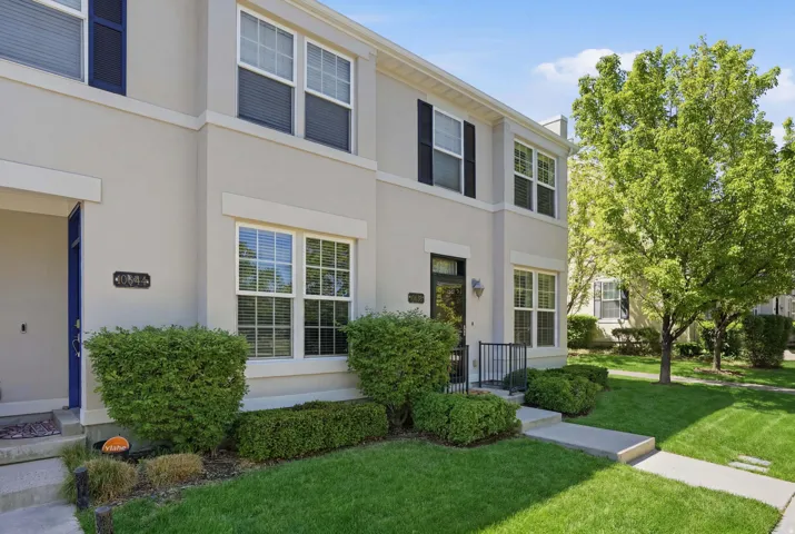 Two-story townhouse featuring a light-colored stucco exterior, multiple windows with white frames, black exterior shutters, a black front door with a glass panel, and a small black metal railing