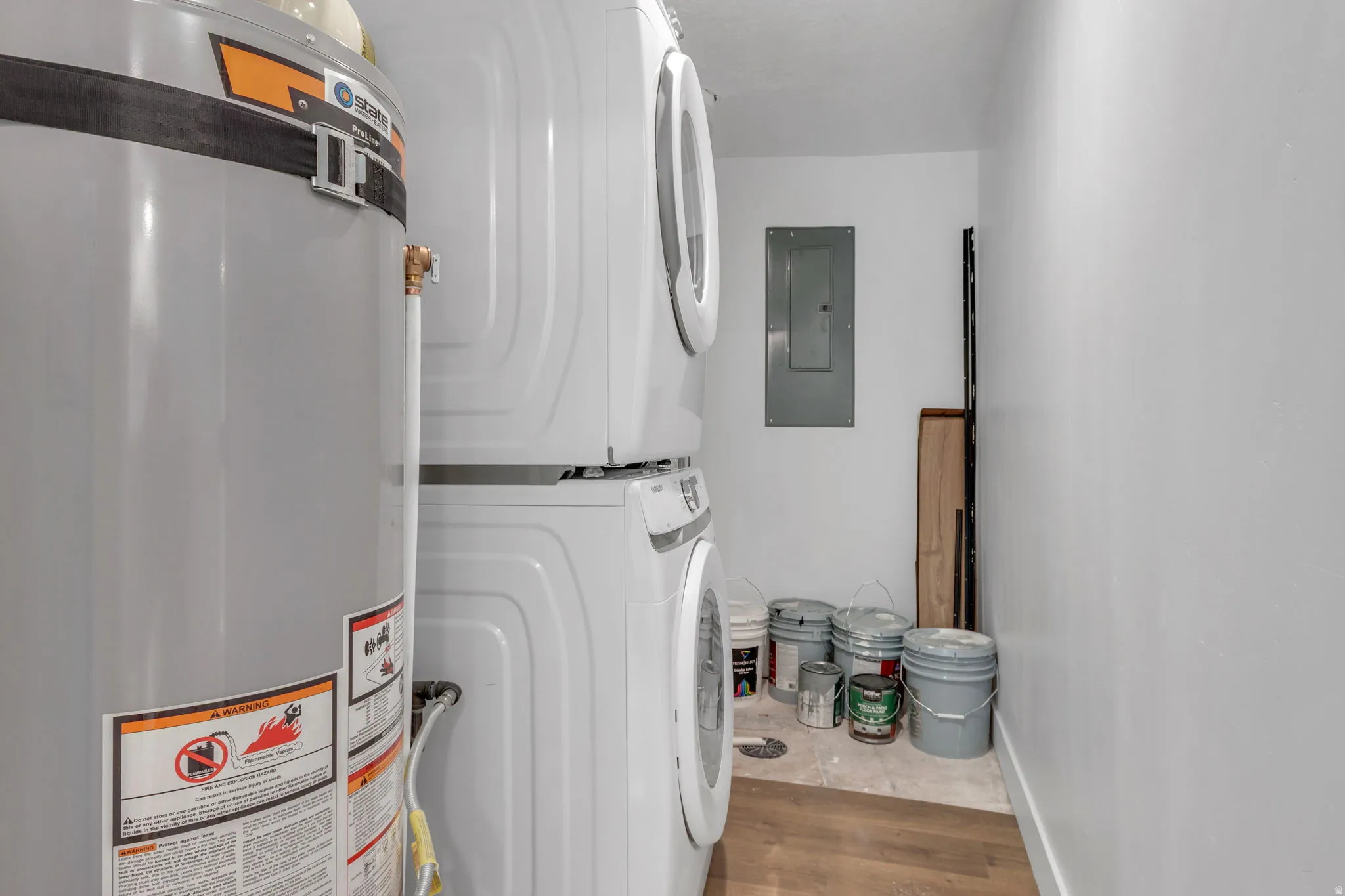 Laundry room featuring secured water heater, electric panel, stacked washer / drying machine, and wood LVP floors