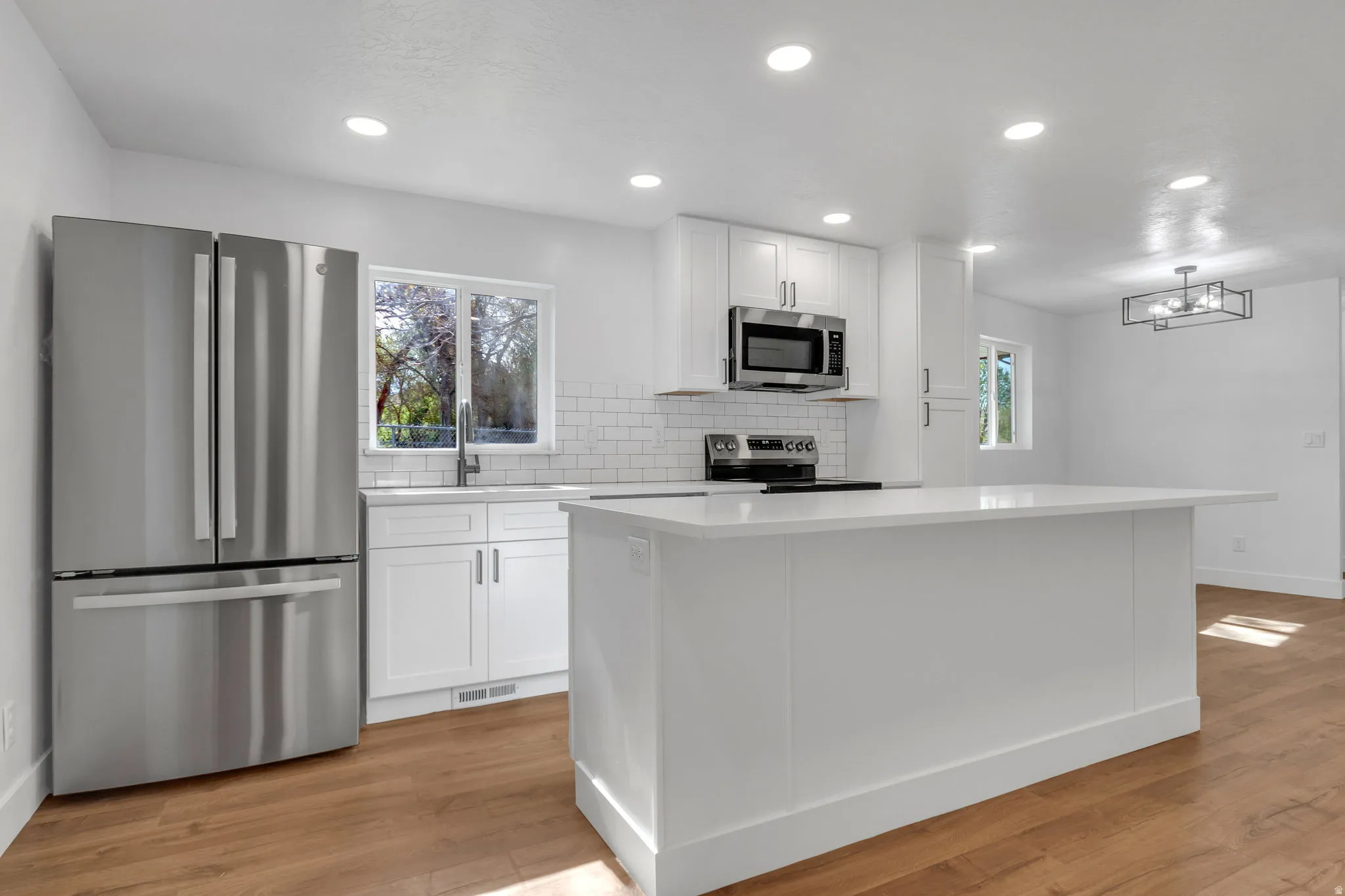 Kitchen featuring stainless steel appliances, white shaker cabinets, tasteful backsplash, quartz island, light wood LVP flooring, and recessed lighting.