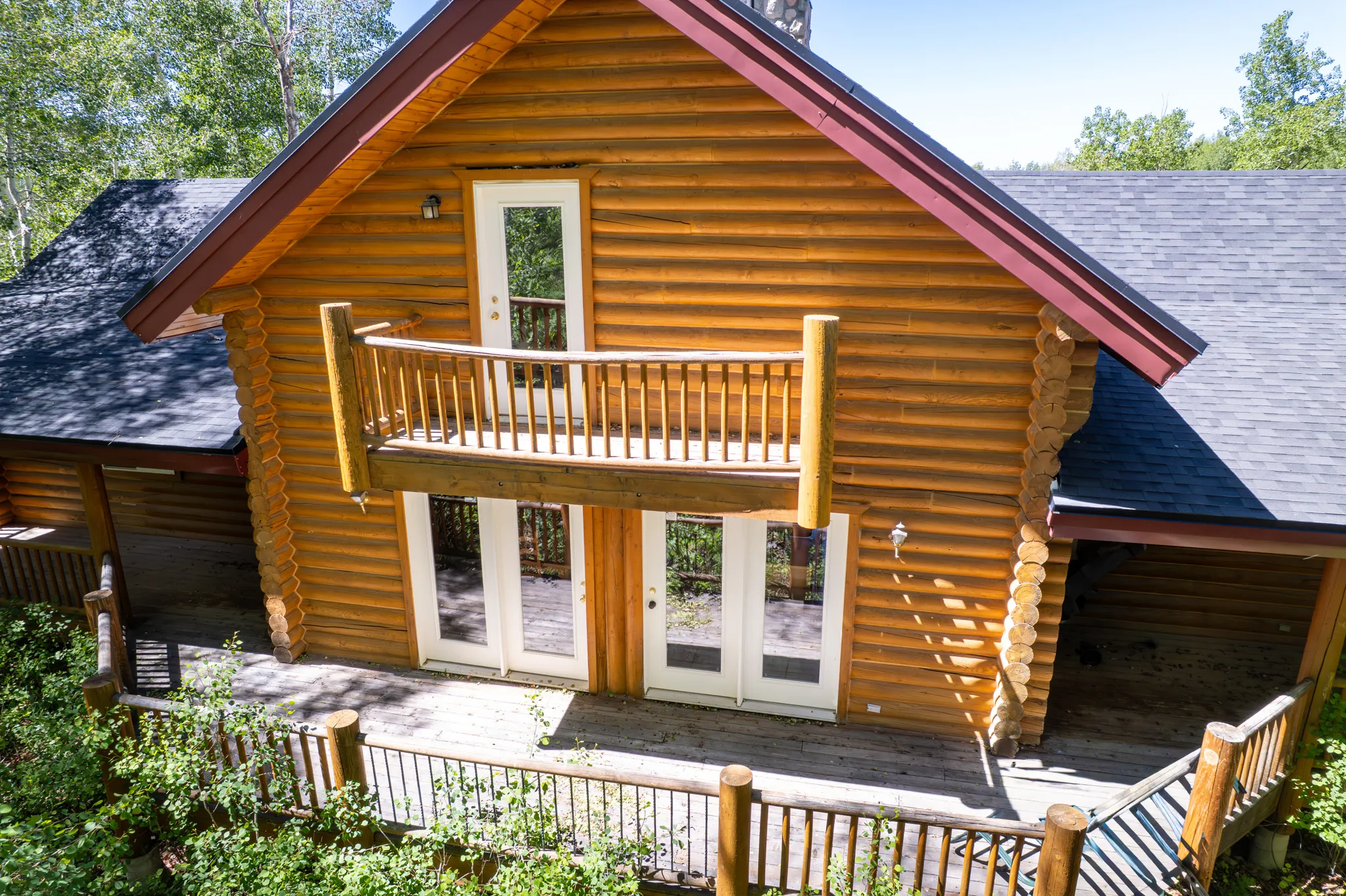 Log home exterior featuring a second-story balcony with wood railing, dual glass-paned doors on the lower level, a dark shingle roof, and a wood deck with railings