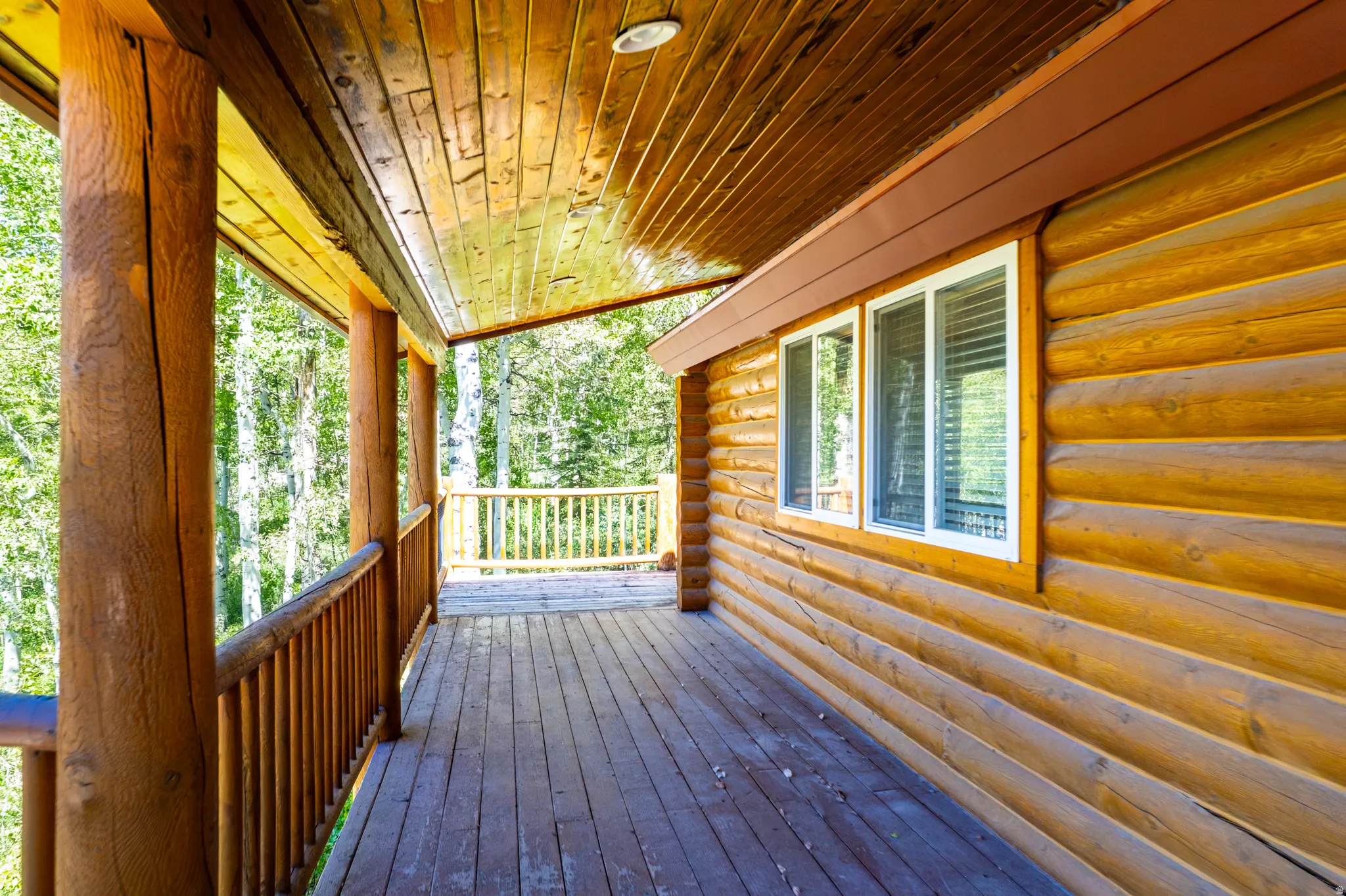 Expansive log cabin porch featuring a wood-plank floor, robust log columns, a wooden railing, and a log exterior wall with white-framed windows