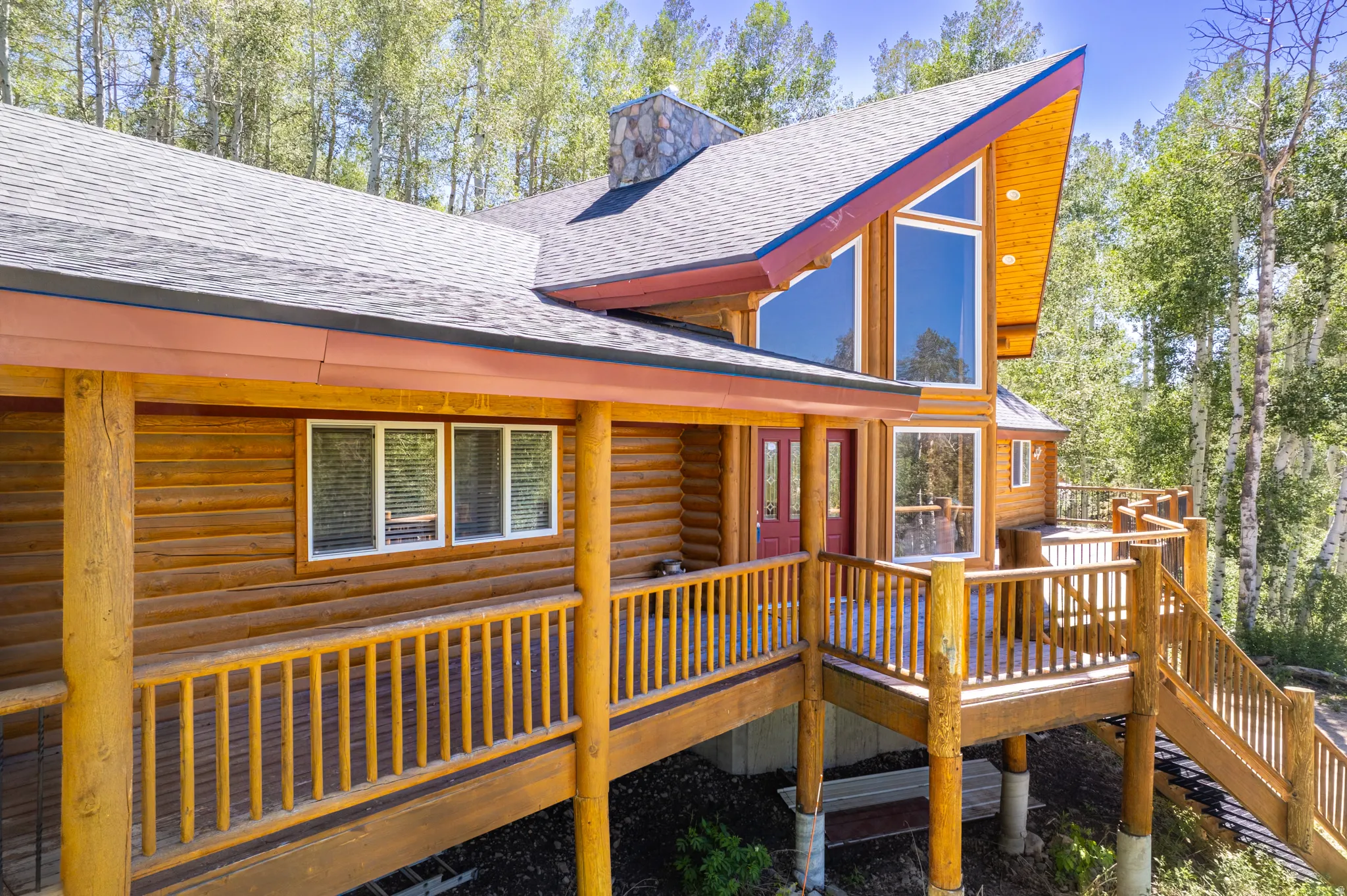 Log cabin exterior with a multi-level deck, pitched roofline, large picture windows, red entry doors, and a stone chimney