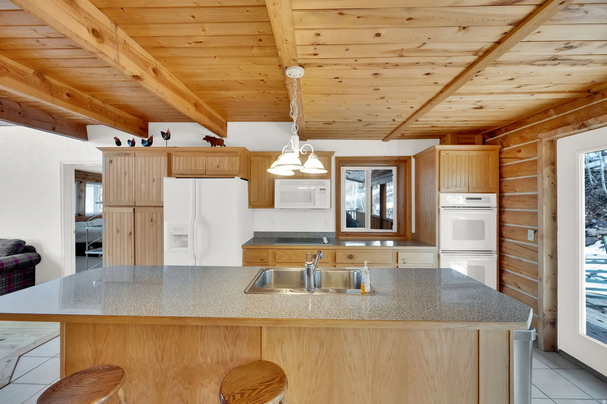 Kitchen featuring light tile patterned flooring, wooden ceiling, white appliances, light wood finish cabinetry, and a kitchen island with sink