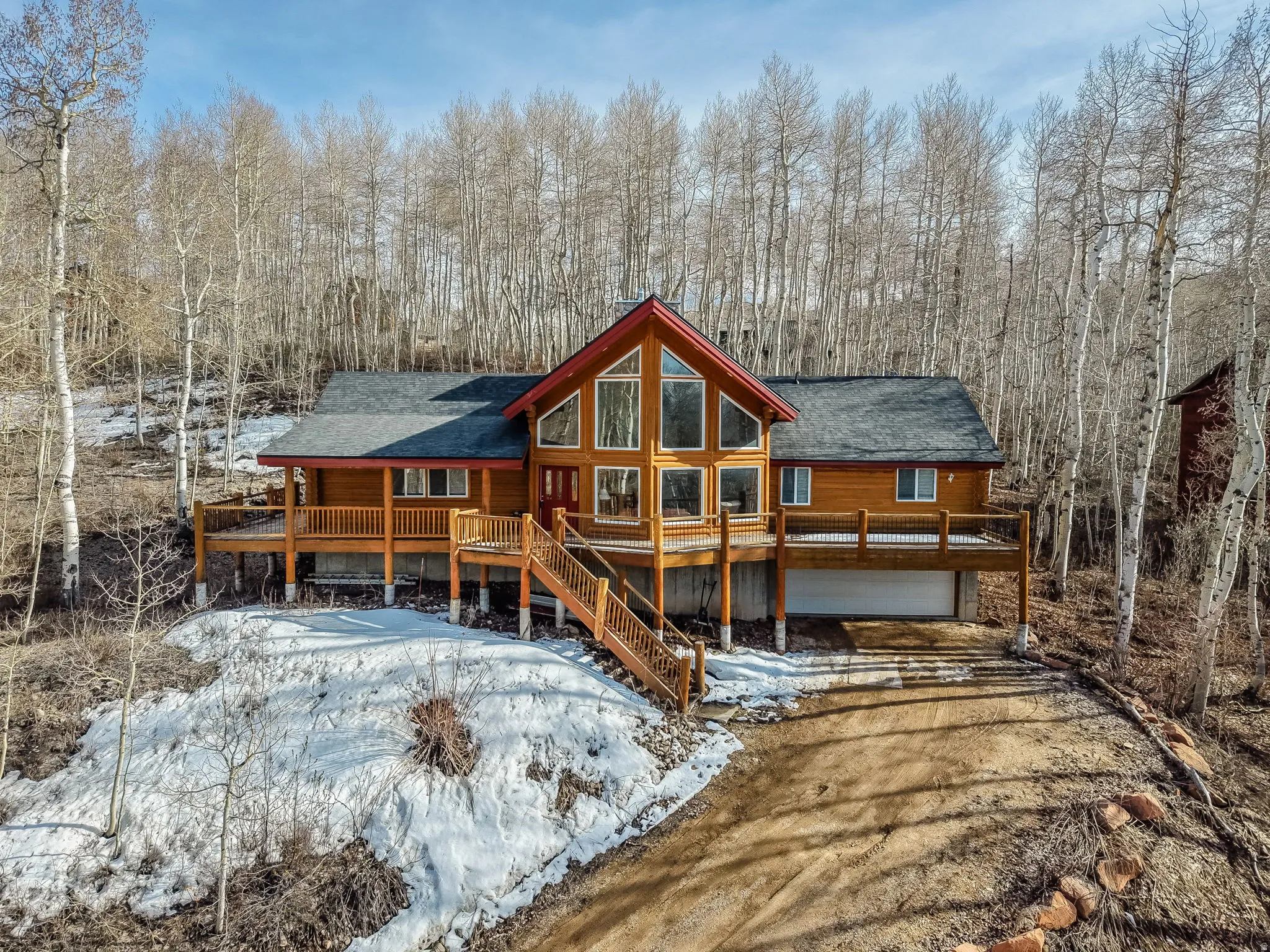 Snow covered back of property featuring an attached garage, driveway, a shingled roof, and a wooden deck