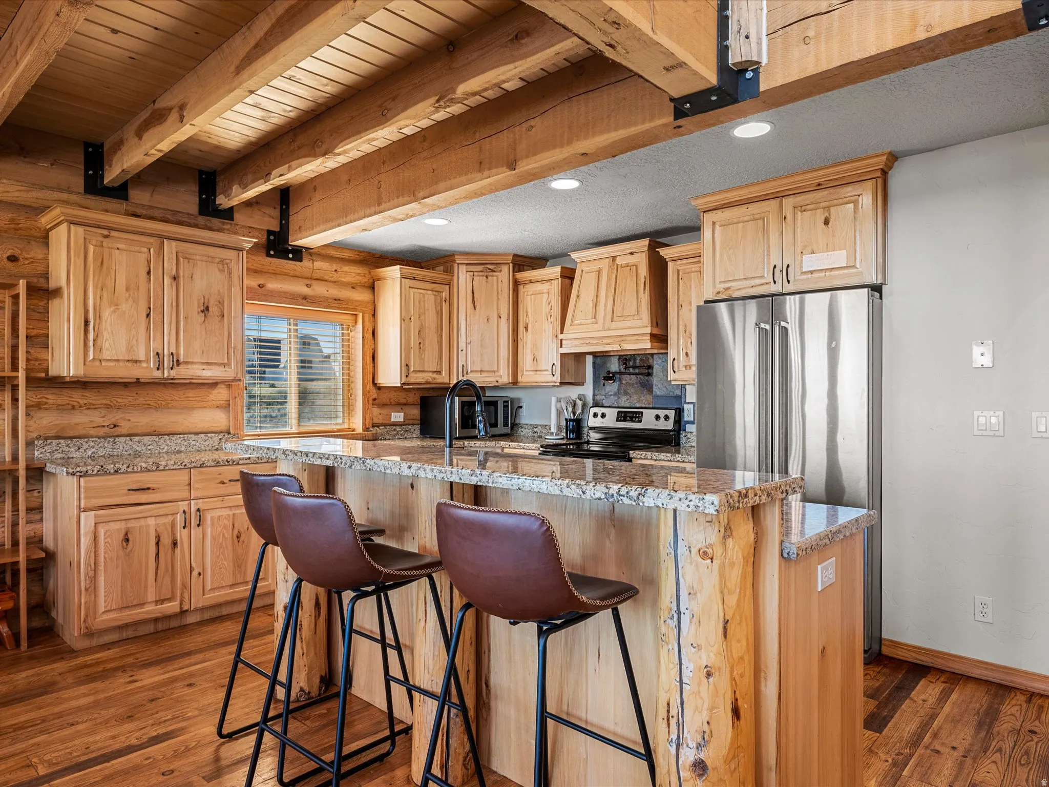 Kitchen with light wood finish cabinetry, dark wood-style flooring, rustic walls, stainless steel appliances, and a center island with sink