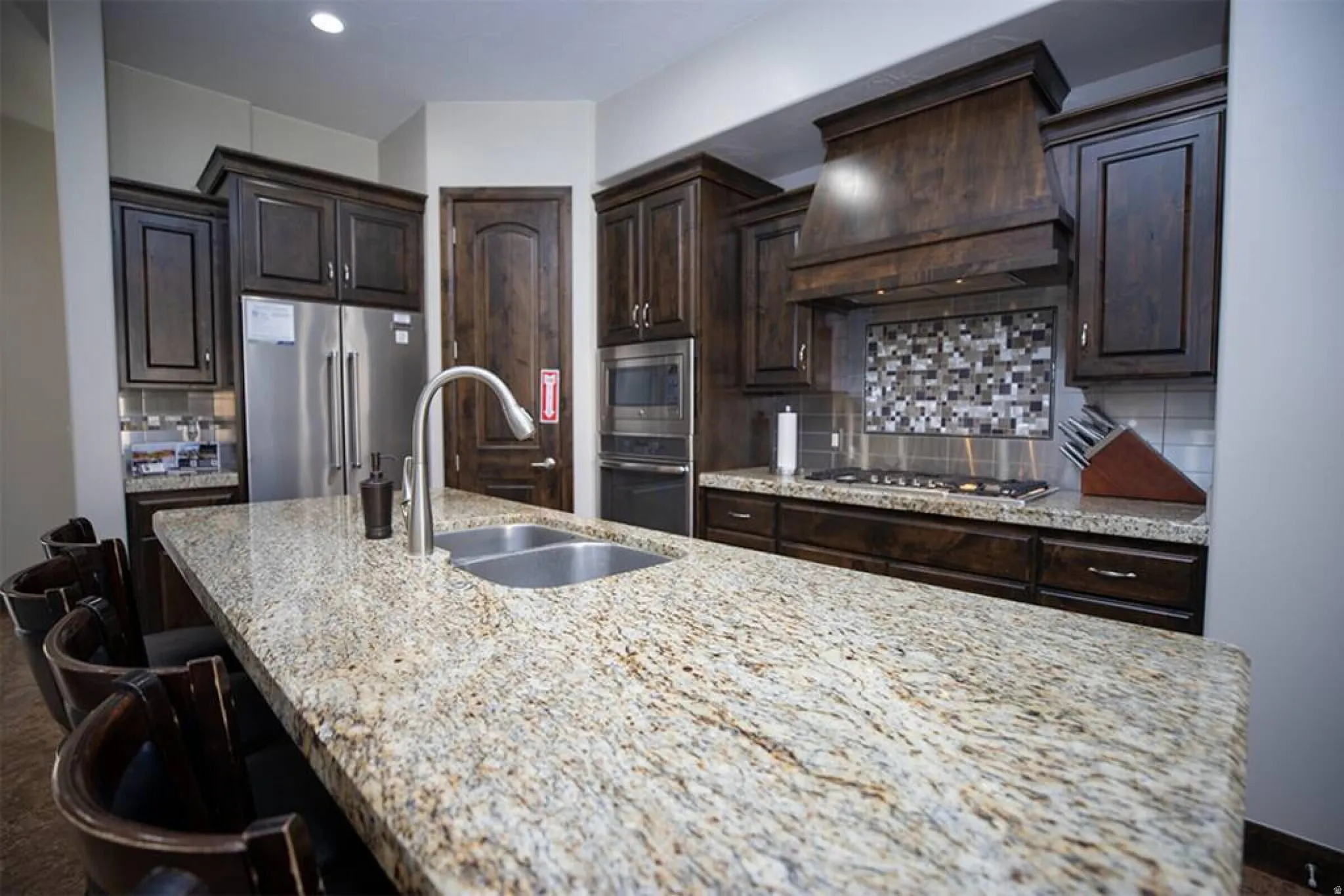 Kitchen with dark wood finish cabinets, stainless steel appliances, a large island with sink, and light stone counters