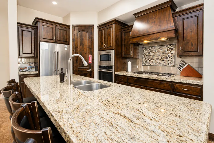 Spacious kitchen island with an undermount double basin stainless steel sink, dark wood cabinetry, a mosaic tile backsplash, a stainless steel gas cooktop, and built-in stainless steel appliances