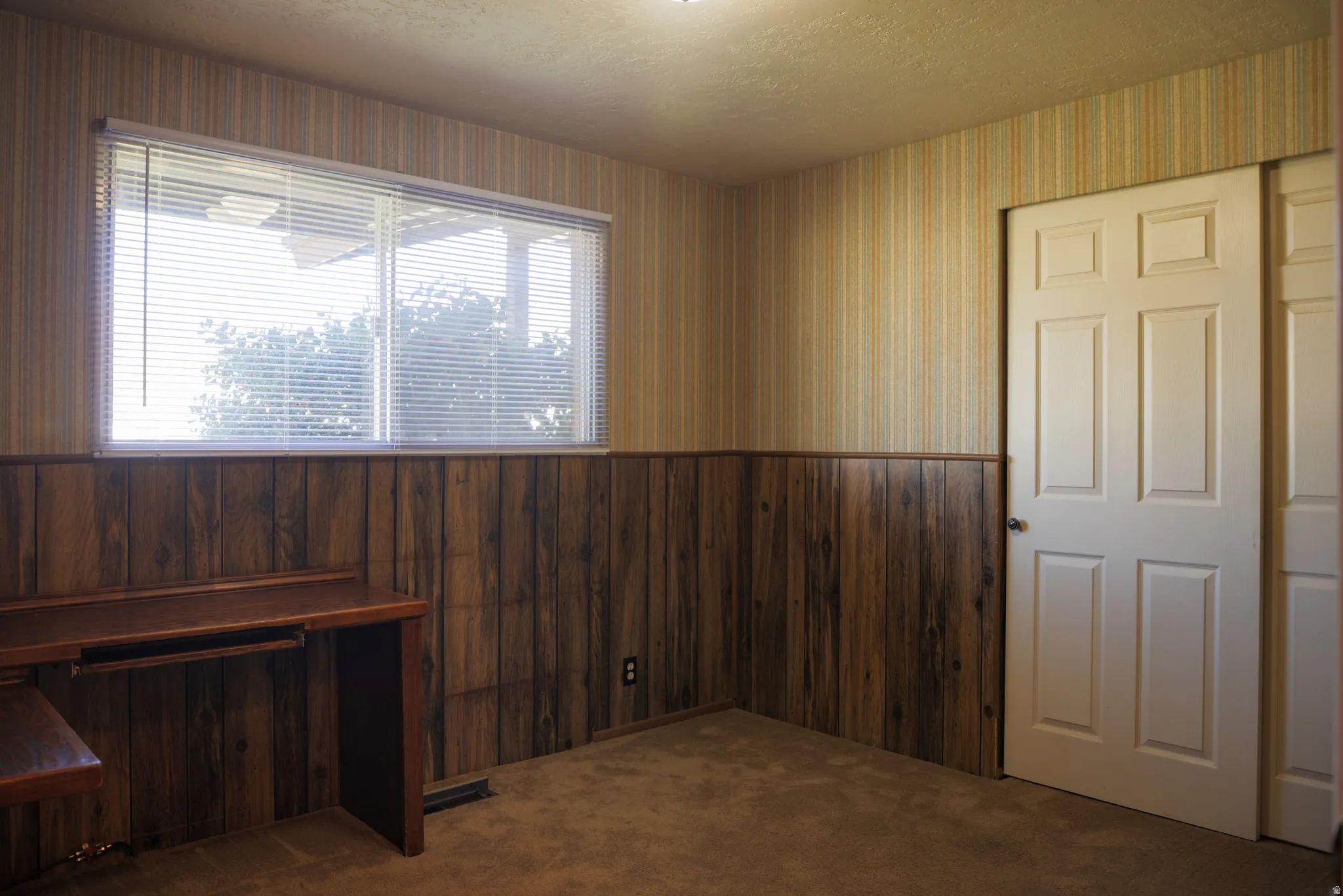 Empty room with a textured ceiling, dark colored carpet, wooden walls, wallpapered walls, and wainscoting