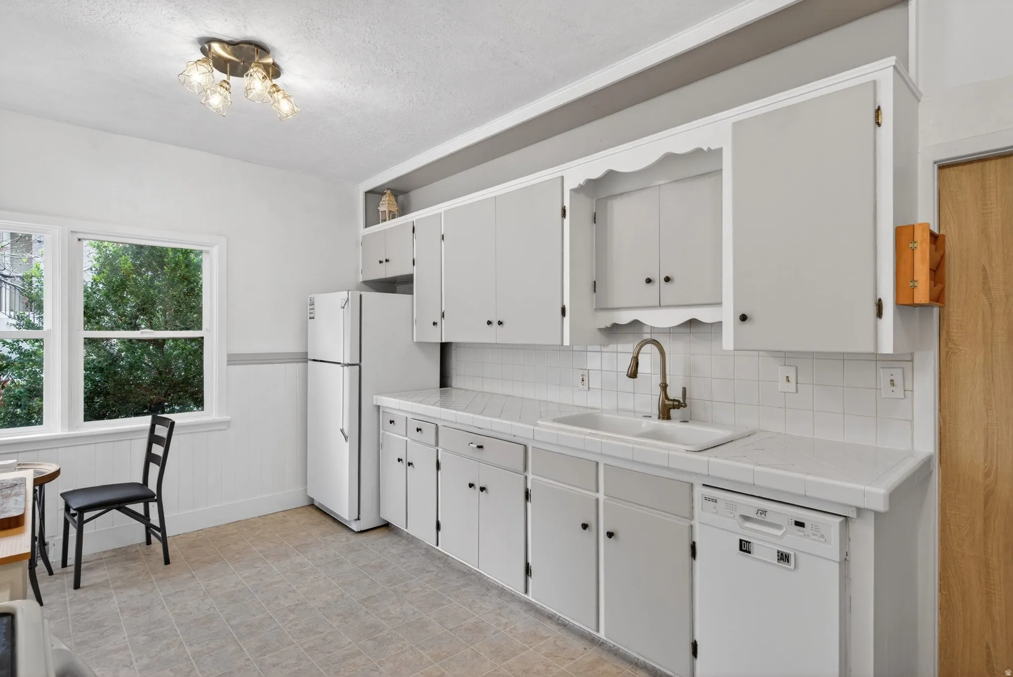 Kitchen with tile counters, white appliances, a wainscoted wall, white cabinetry, and a textured ceiling
