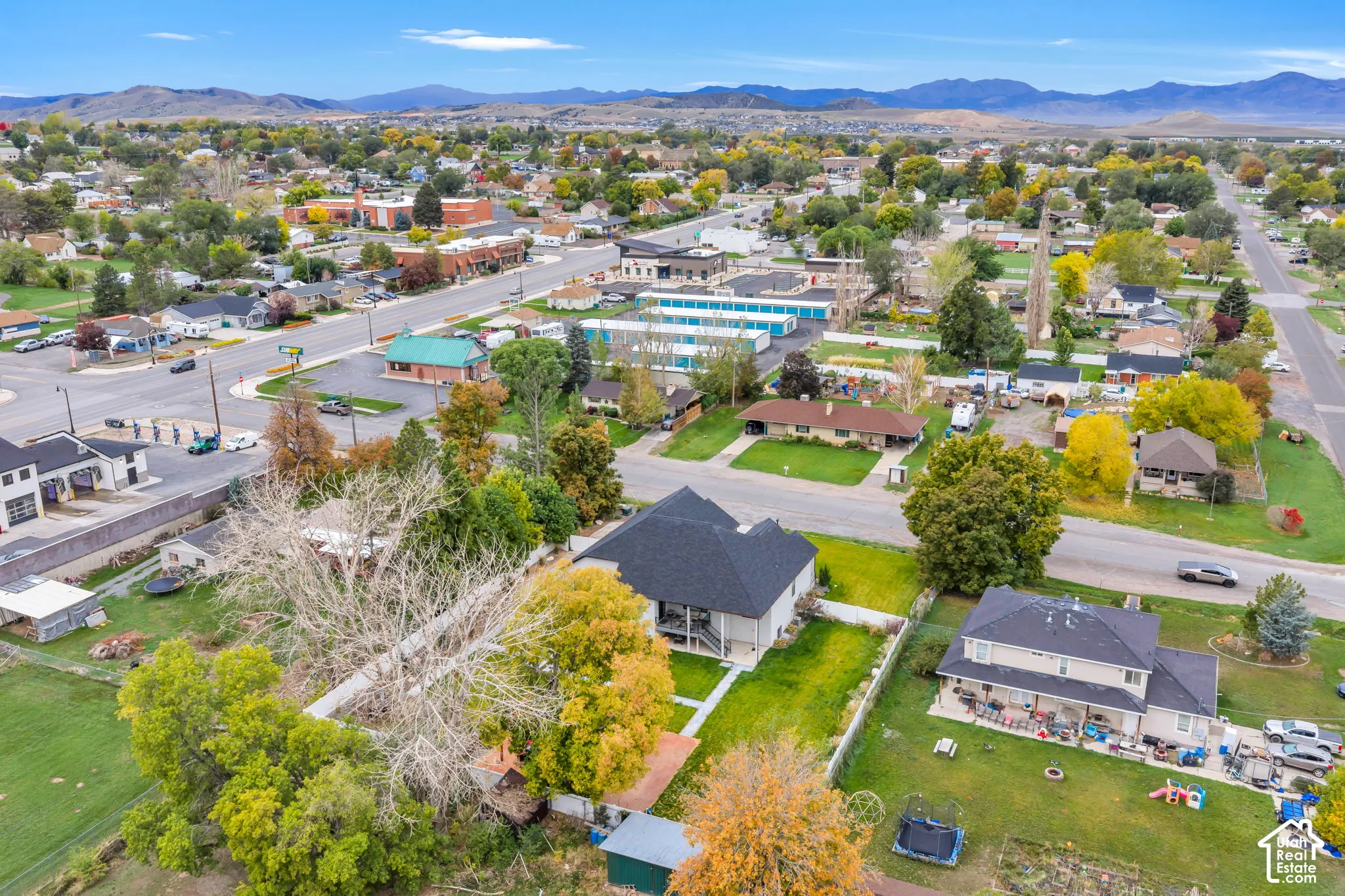 Aerial perspective featuring a multi-story residence with a dark shingled roof, manicured lawn, mature trees, and mountain backdrop