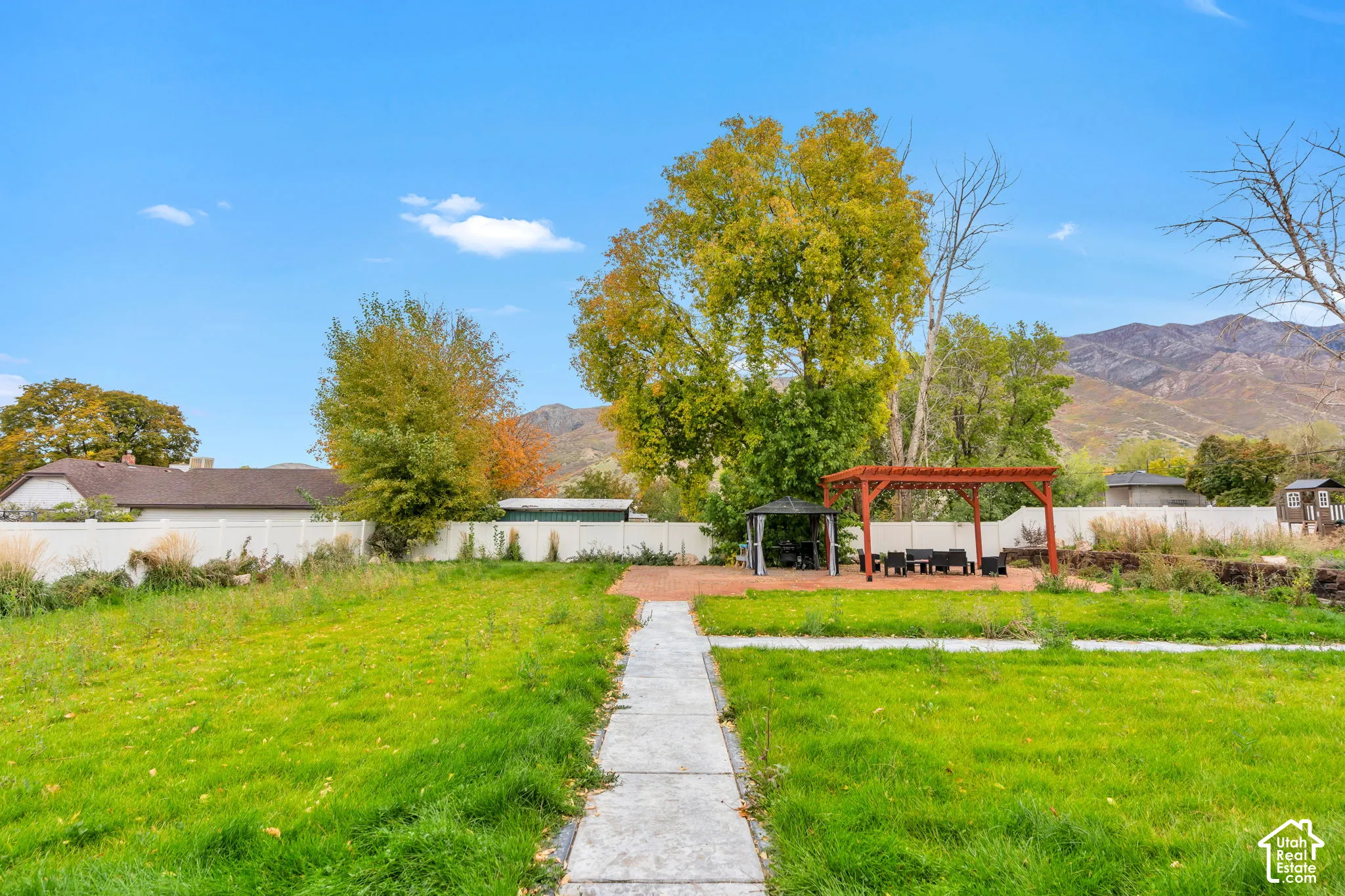 Backyard featuring a paved stone walkway, a wood-frame pergola, a brick patio, a white vinyl perimeter fence, and a mountain backdrop