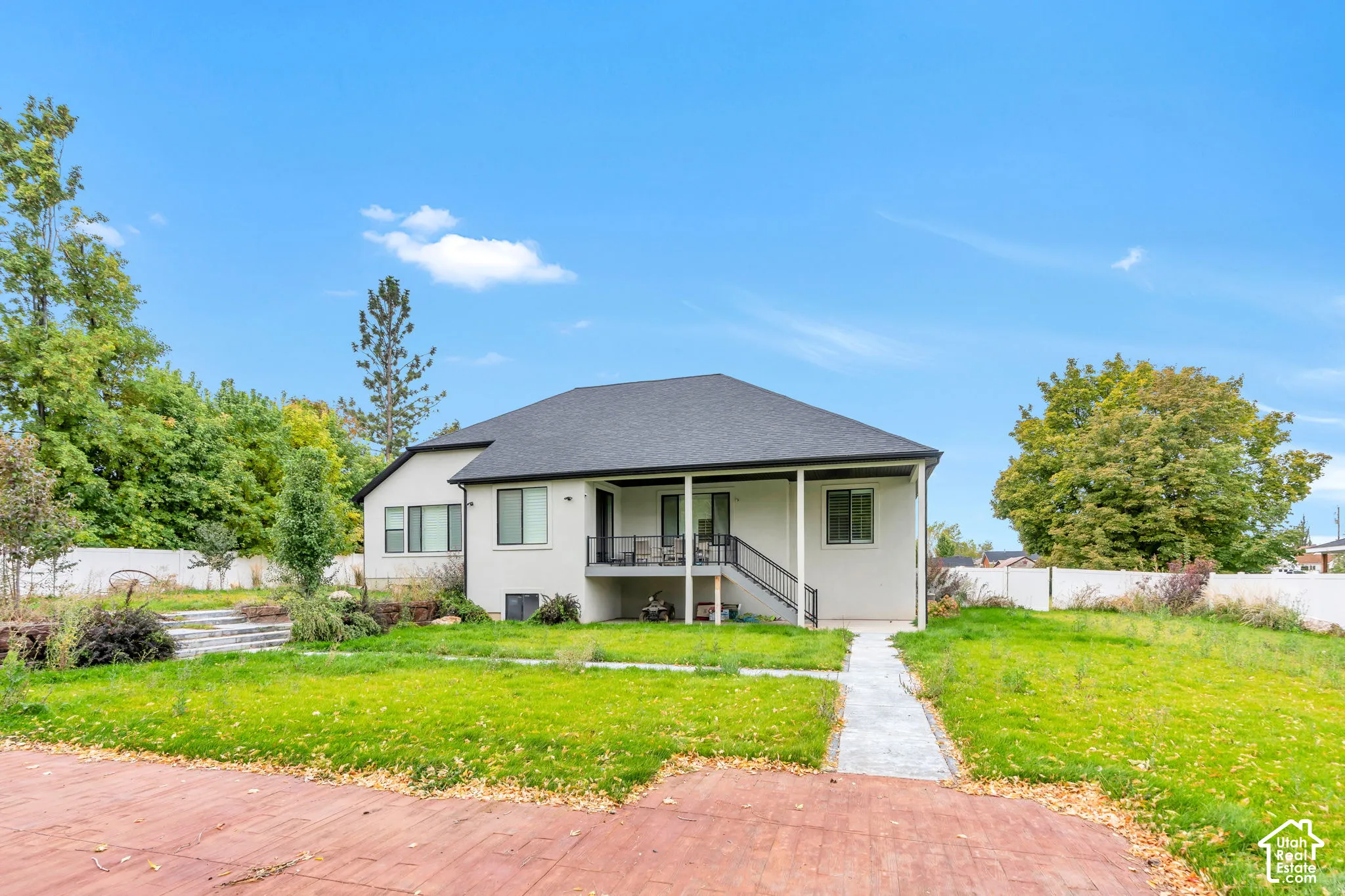 Rear exterior featuring a covered deck with black metal railing, concrete patio, concrete walkway, and white vinyl perimeter fencing
