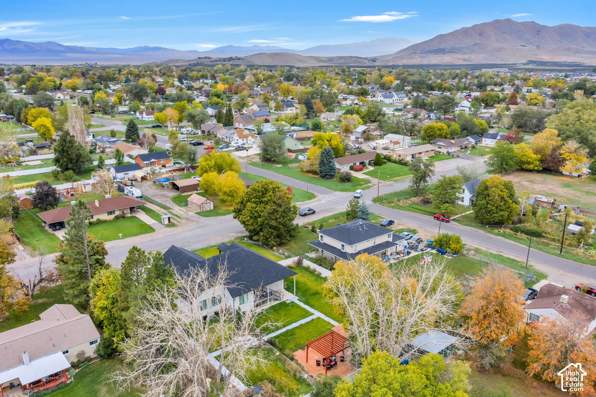 Aerial perspective featuring a multi-level residence with dark shingle roofing, expansive green lawn, mature trees, wood-frame pergola, and mountain backdrop
