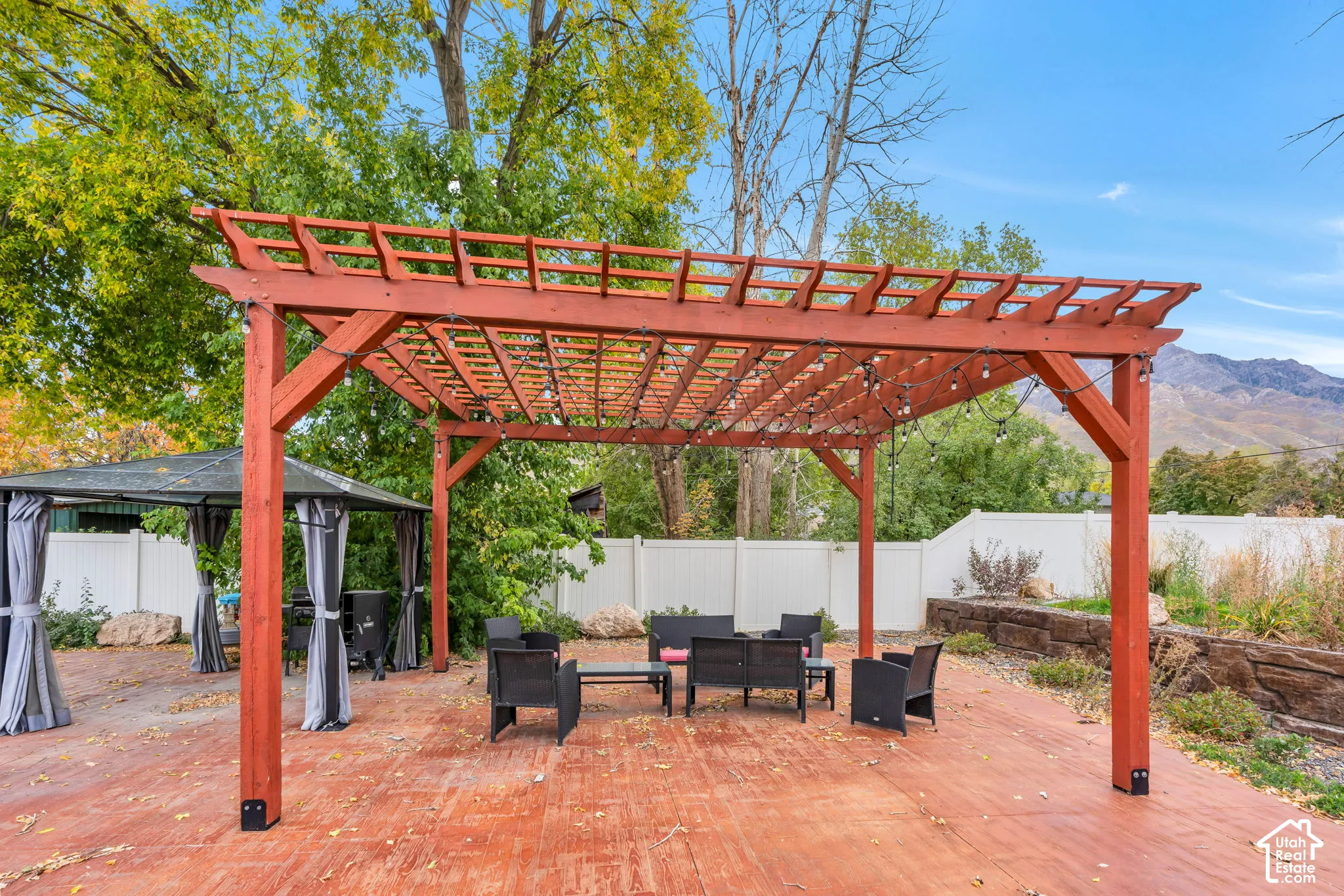 Stained wood pergola structure on a paved patio with a stone retaining wall, enclosed by white vinyl fencing and mountain backdrop