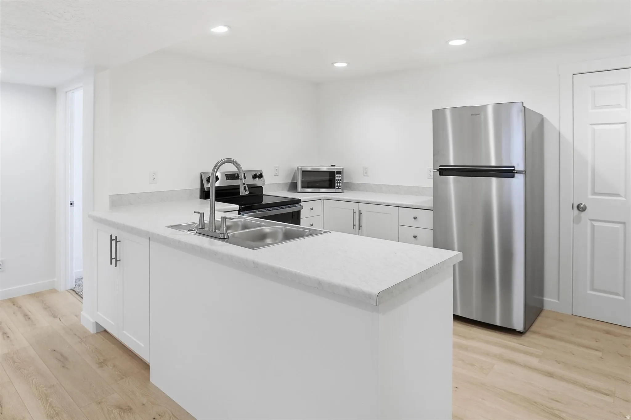 Basement kitchen with stainless steel appliances, a peninsula, white cabinets, light wood finished floors, and light countertops