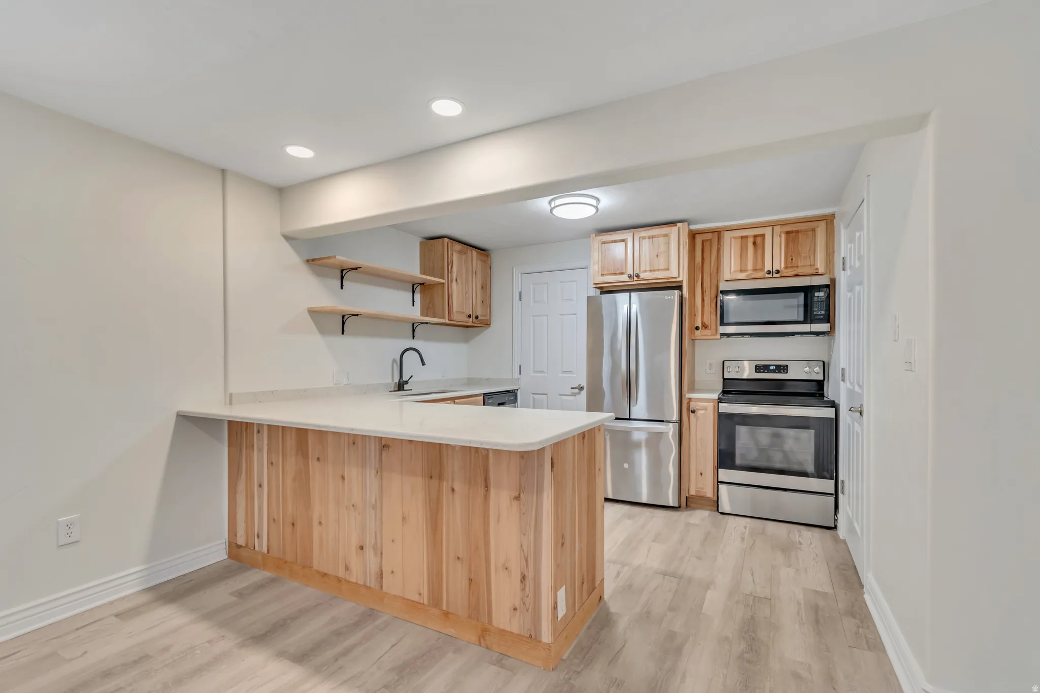 Kitchen with stainless steel appliances, a peninsula, light countertops, open shelves, and light wood finish cabinetry