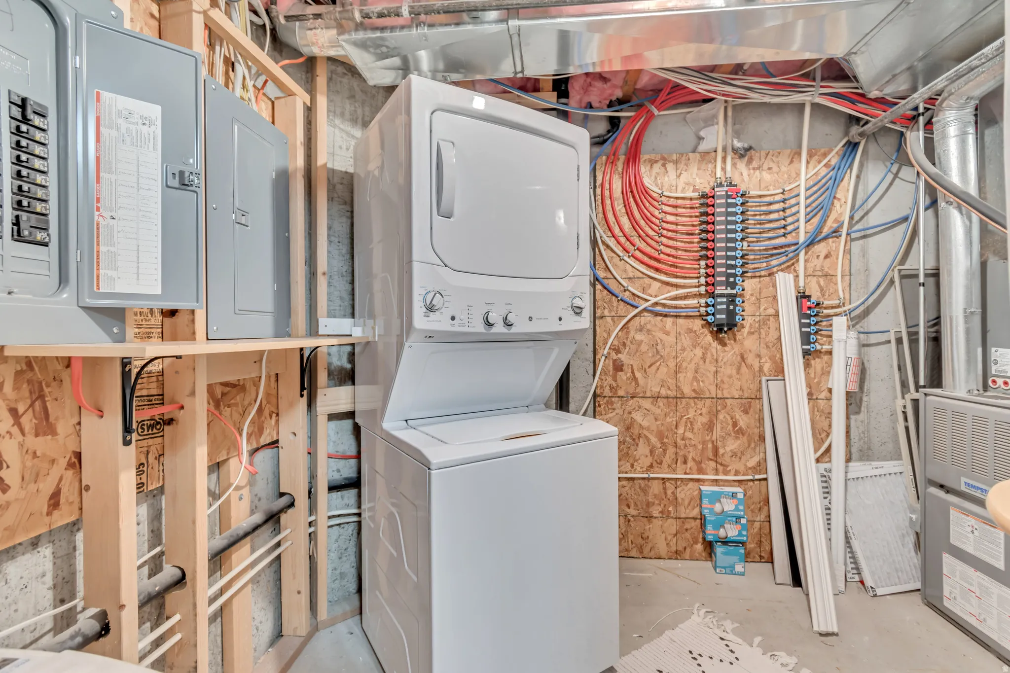 Laundry area featuring electric panel, stacked washer / drying machine, unfinished concrete floors, and heating unit