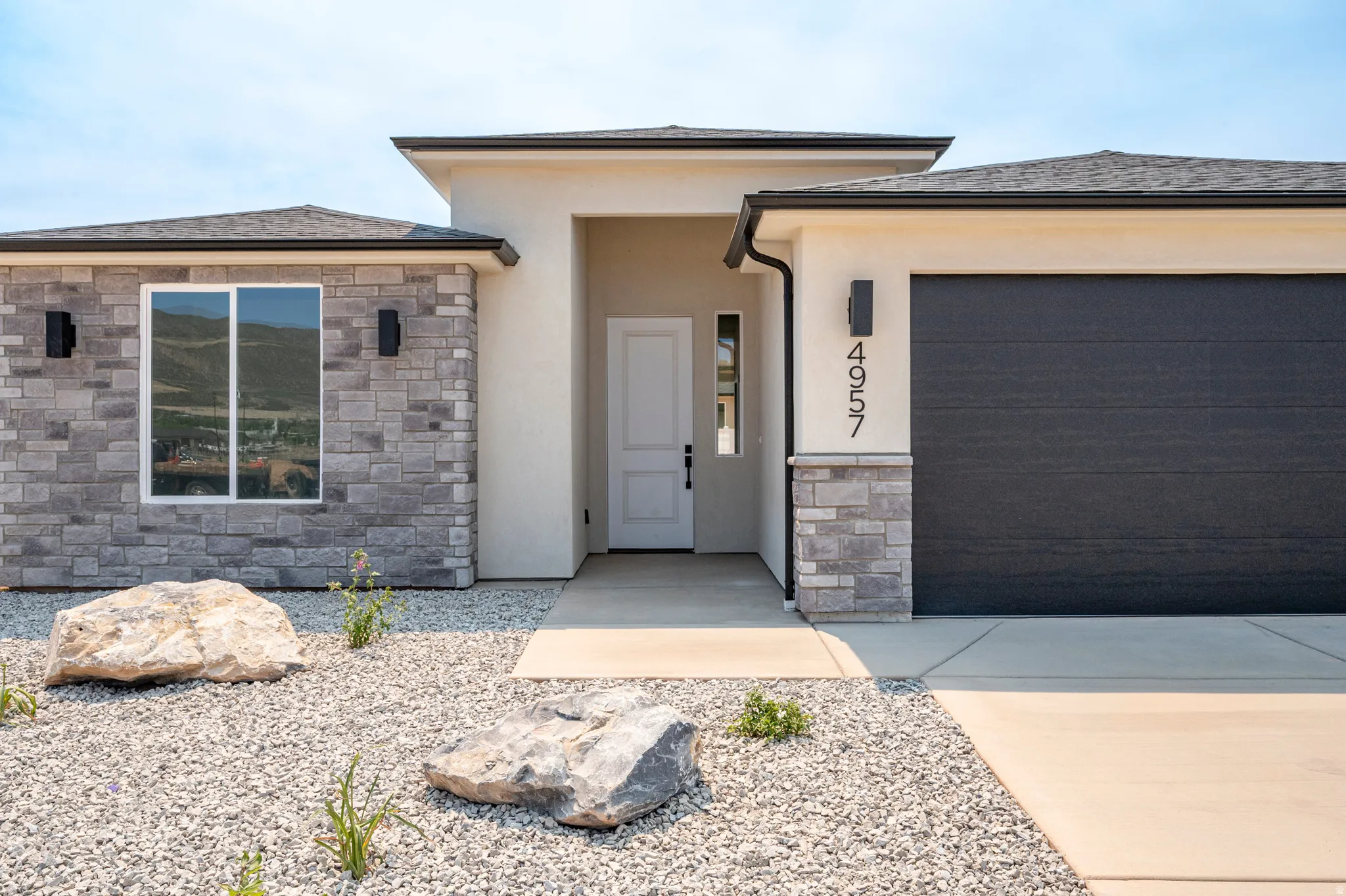Contemporary stucco and stone exterior featuring a dark gray garage door, white entry door, and dark exterior lighting fixtures
