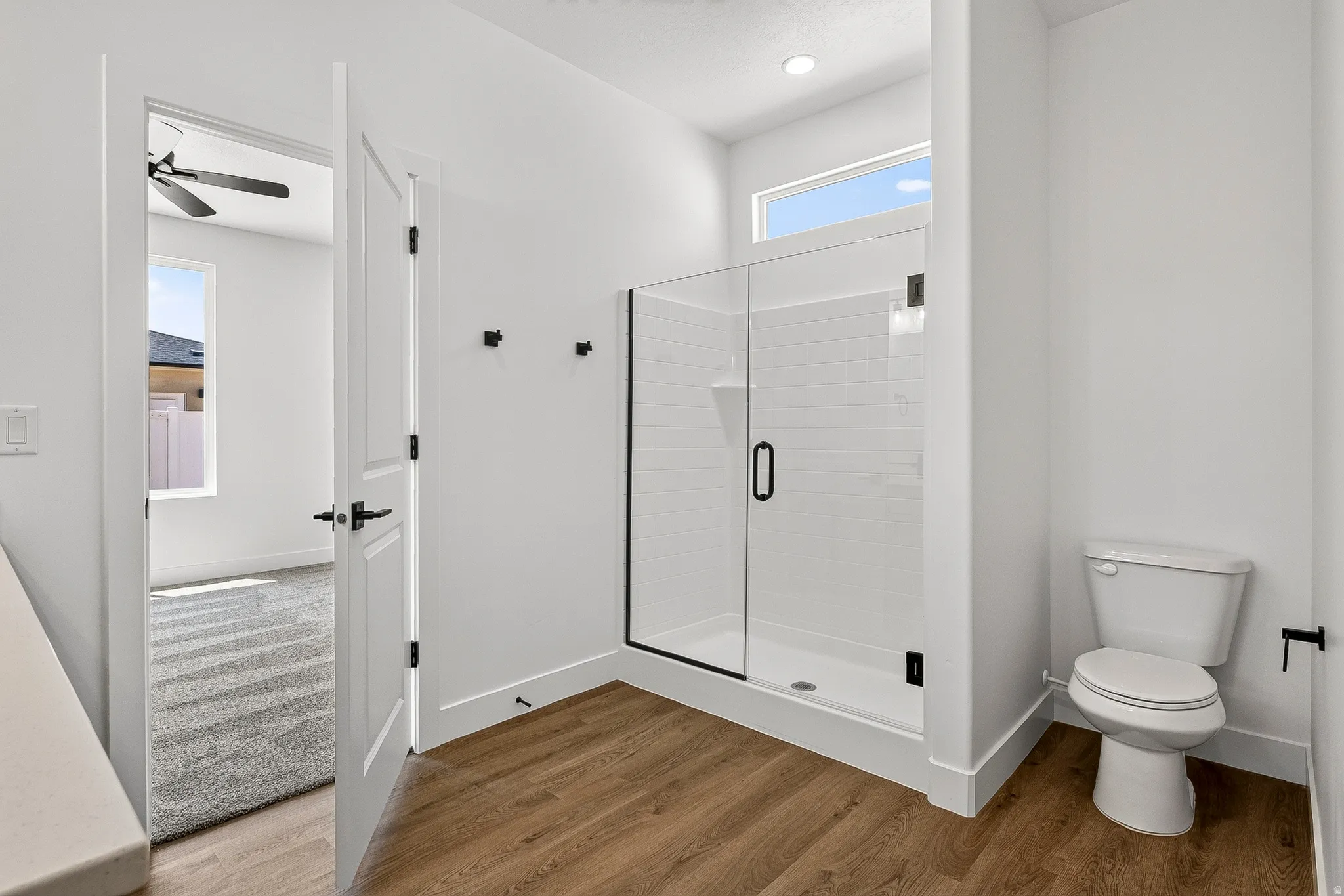 Bathroom featuring wood-finish flooring, a glass-enclosed shower with subway tile, a white toilet, and a high transom window