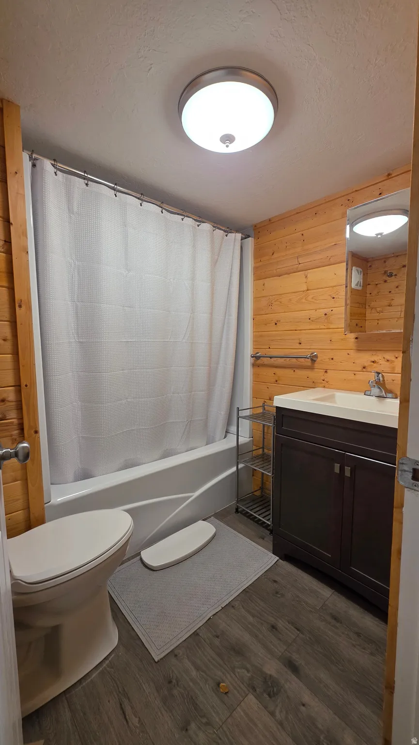 Bathroom featuring vanity, wood walls, a textured ceiling, shower / bathtub combination with curtain, and dark wood-type flooring
