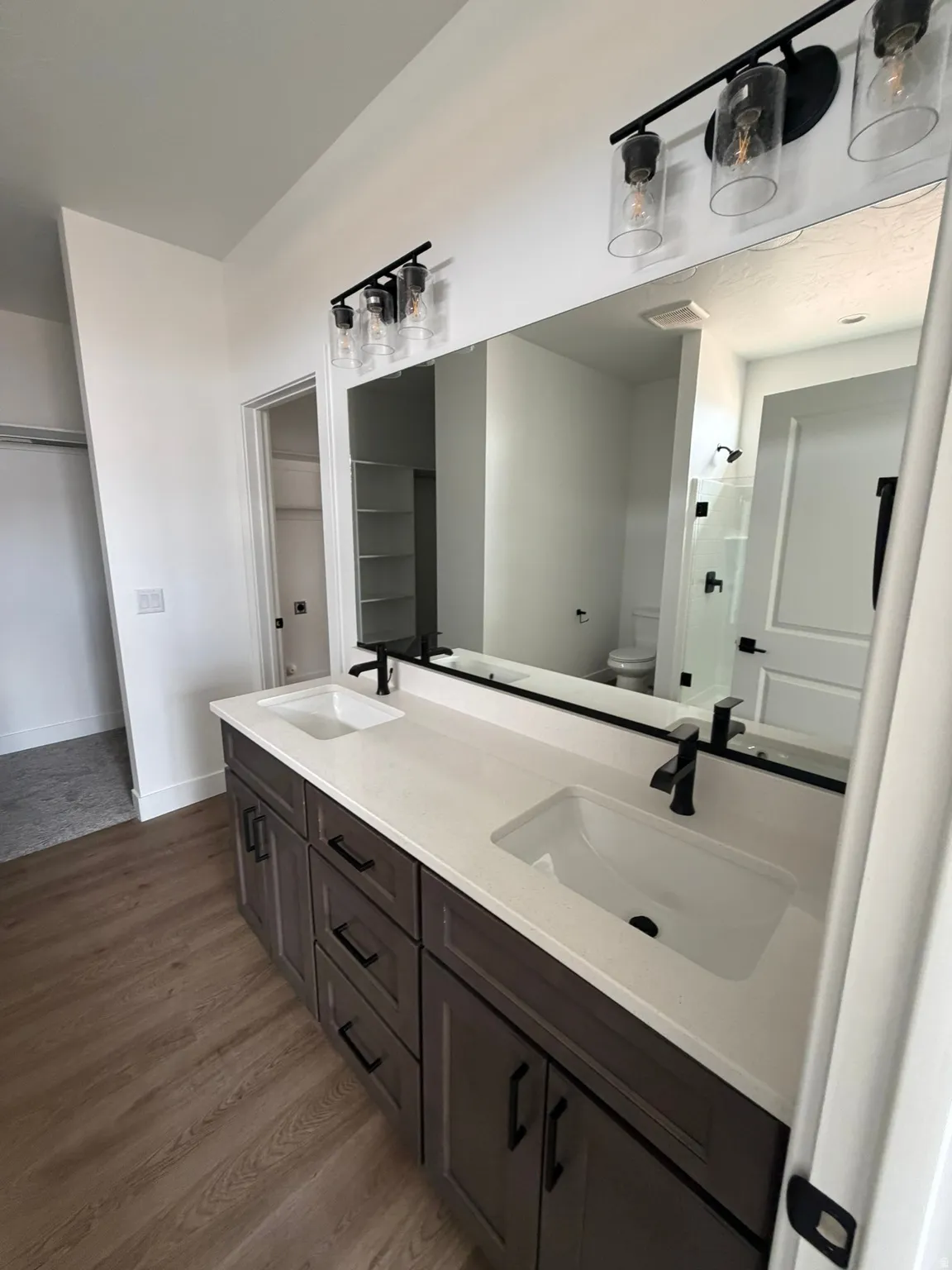Dual vanity with white countertop and integrated sinks, matte black faucets, wood-finish flooring, and overhead lighting fixtures