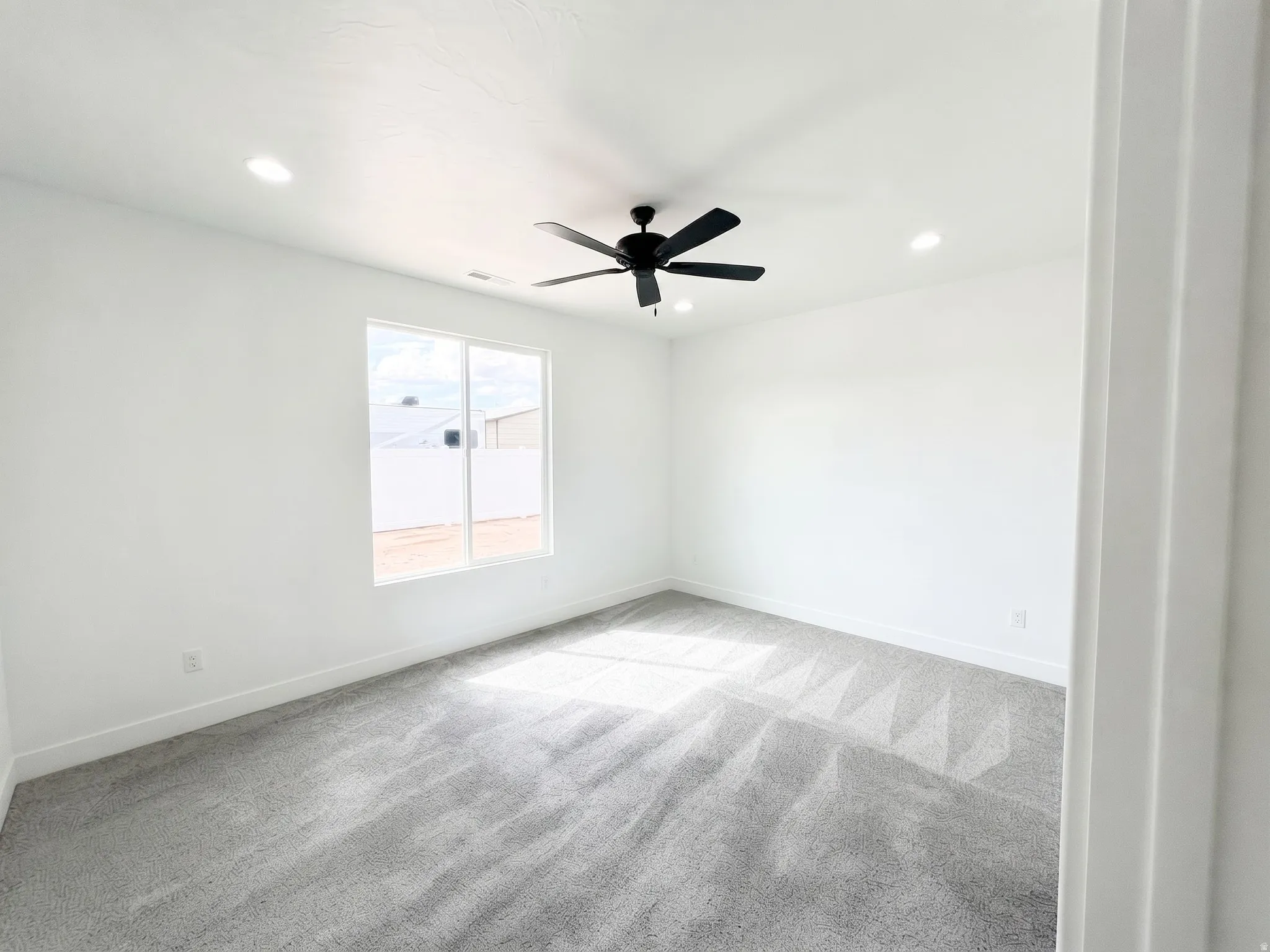 Recessed lighting, ceiling fan, white walls, gray carpeting, and large window
