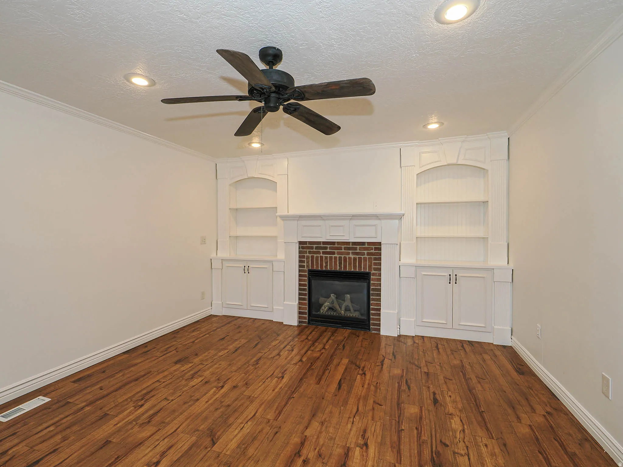 Unfurnished living room featuring built in shelves, dark wood-style floors, ceiling fan, ornamental molding, and a textured ceiling