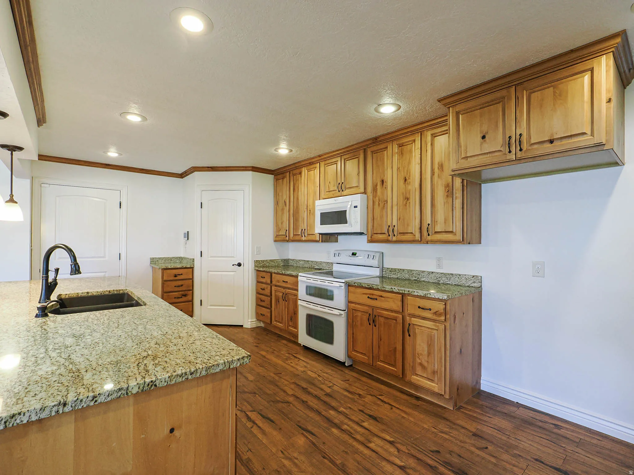 Kitchen featuring wood finish cabinetry, light stone counters, white appliances, dark wood-style floors, and decorative light fixtures