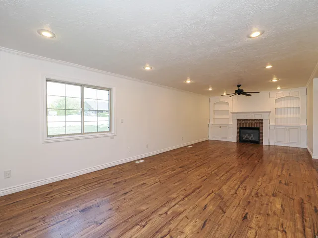 Unfurnished living room featuring a brick fireplace, built in features, dark wood finished floors, a textured ceiling, and ceiling fan