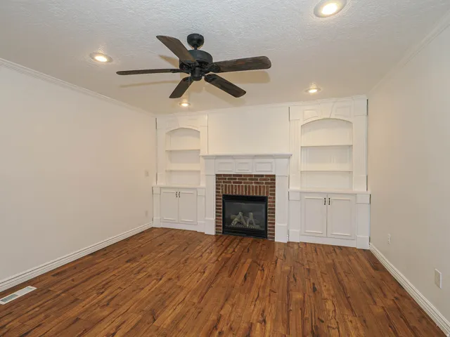Unfurnished living room featuring built in shelves, dark wood-style floors, ceiling fan, ornamental molding, and a textured ceiling