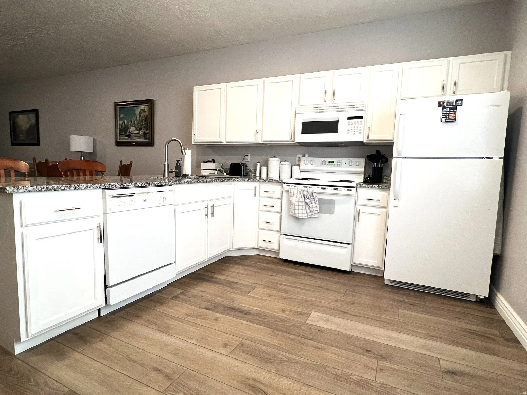 Kitchen with white appliances, white cabinets, light stone countertops, light wood-style flooring, and a textured ceiling