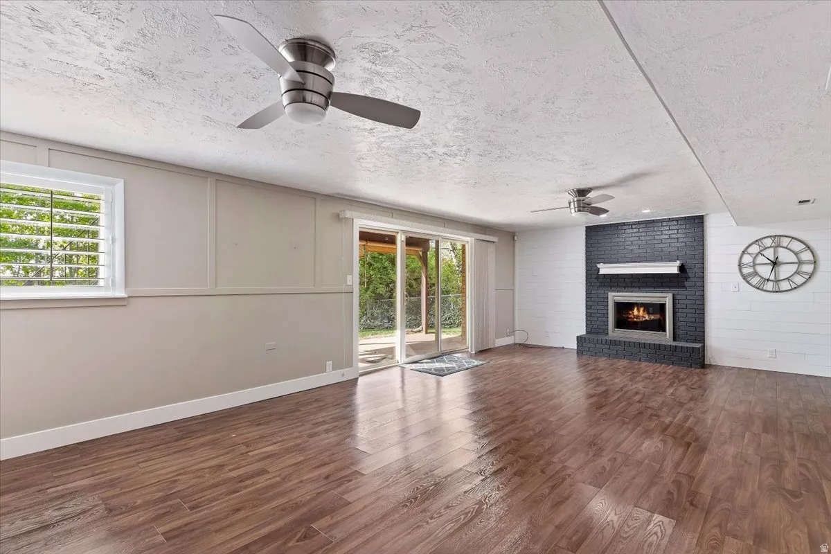 Unfurnished living room featuring a brick fireplace, a textured ceiling, dark wood finished floors, and ceiling fan