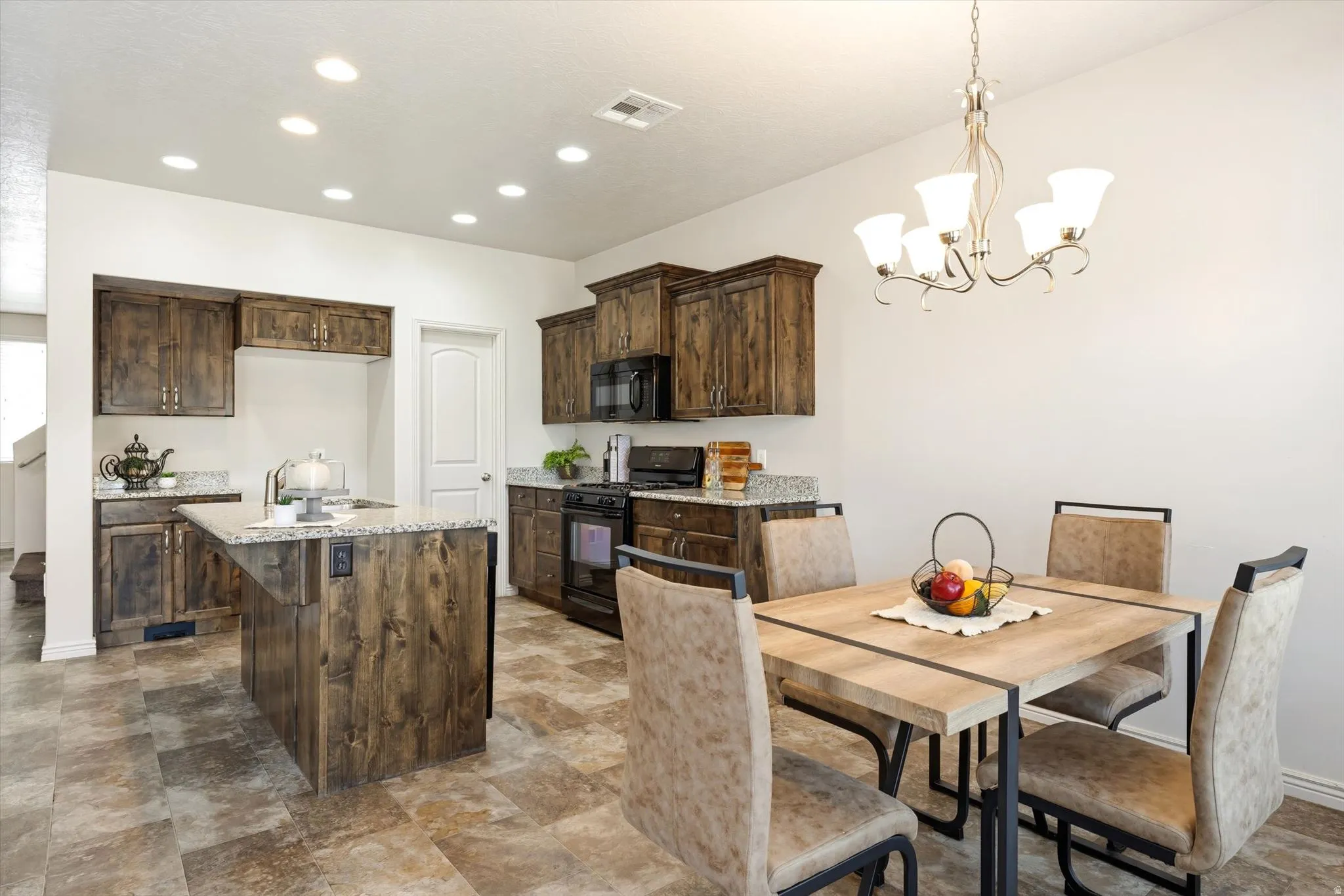 Kitchen with black appliances, dark wood finish cabinetry, a center island with sink, stone finish floors, and hanging lights