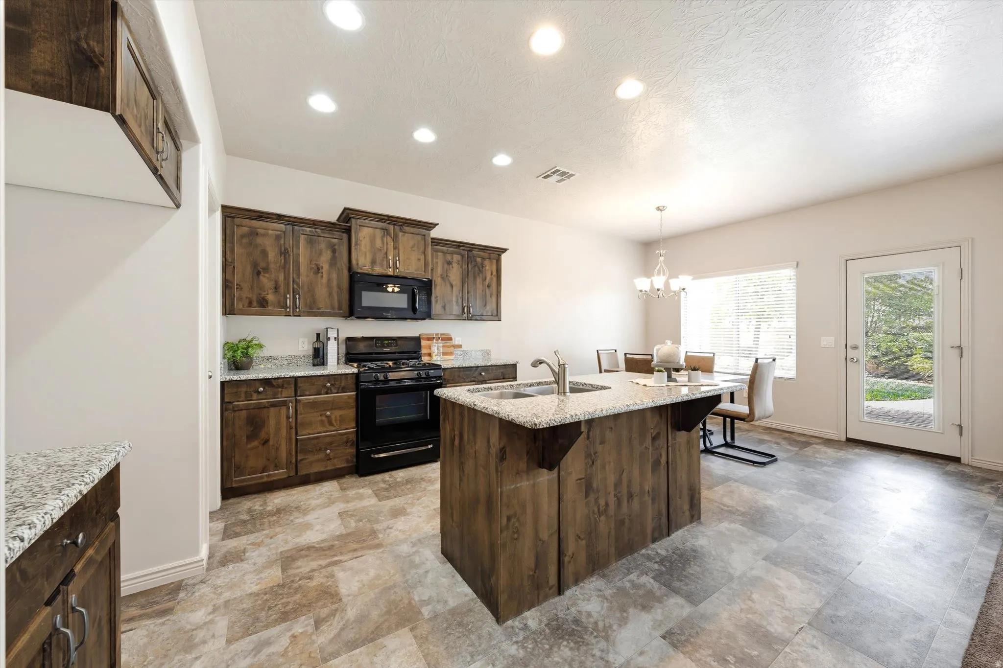 Kitchen with black appliances, a center island with sink, dark wood finish cabinets, stone finish flooring, and light stone countertops