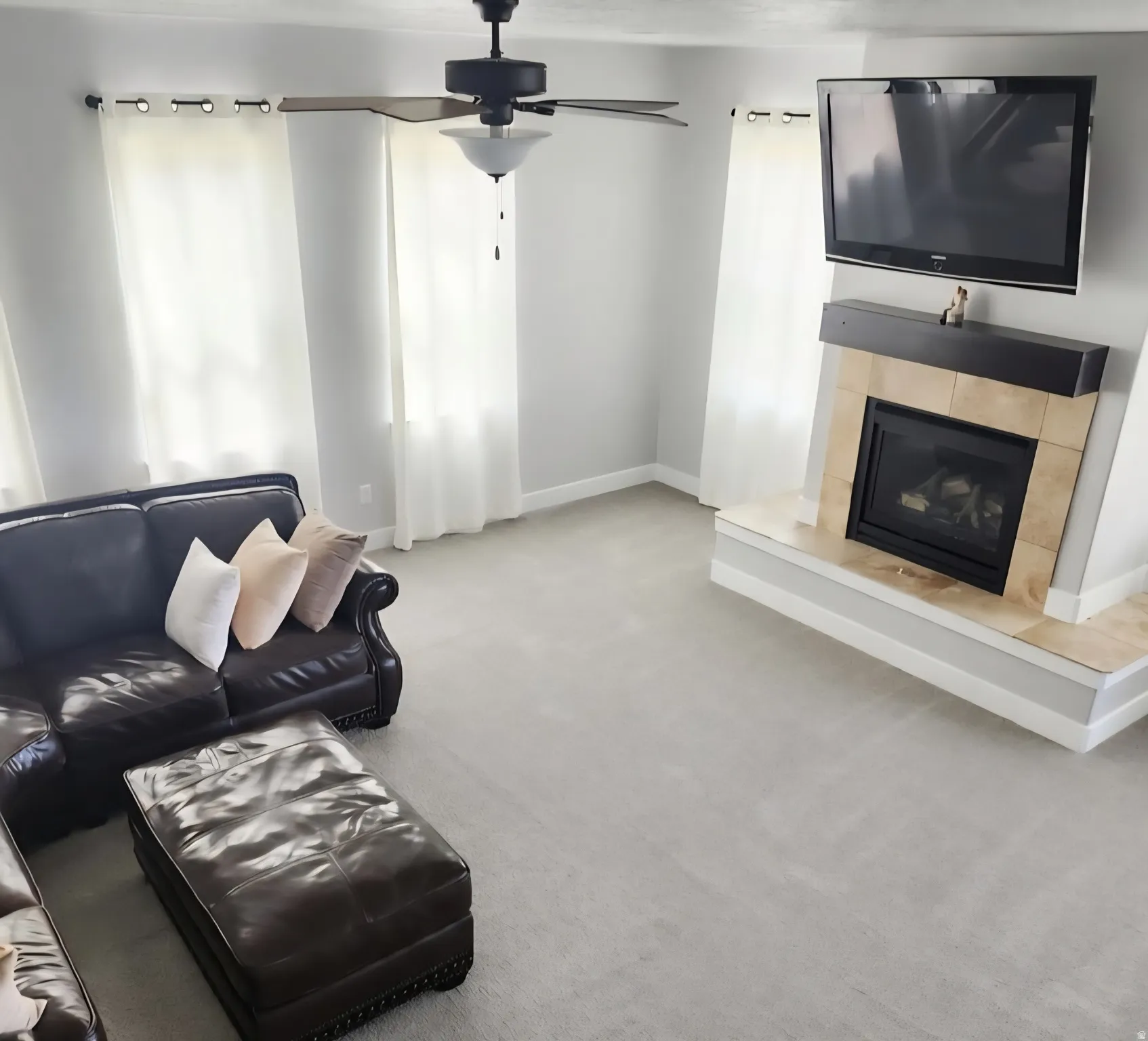 Spacious living area featuring light gray walls, plush carpeting, a ceiling fan with integrated lighting, and multiple windows with white curtain panels