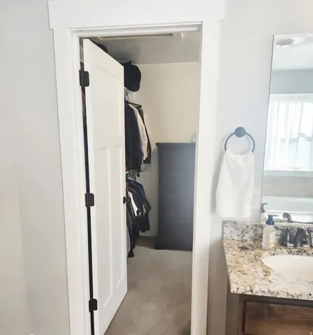 Walk-in closet with carpet flooring and built-in shelving, accessible from a bathroom featuring a granite-style countertop vanity with an undermount sink and dark bronze faucet