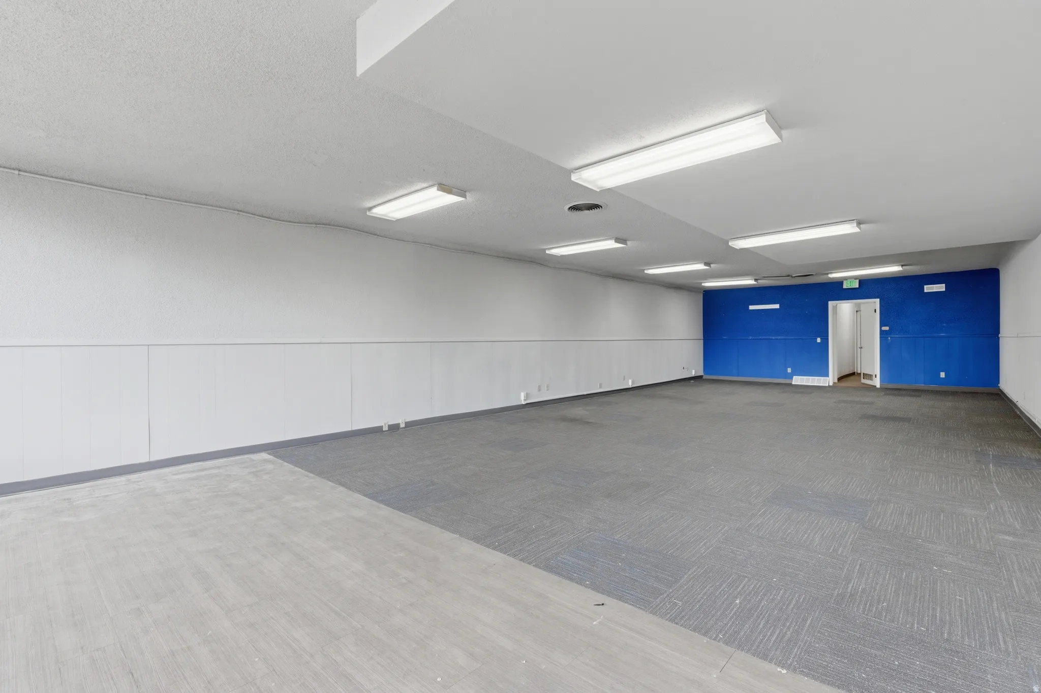 Spacious interior featuring a blue accent wall, white wainscoting, a drop ceiling with fluorescent lighting, and a combination of carpet and concrete flooring