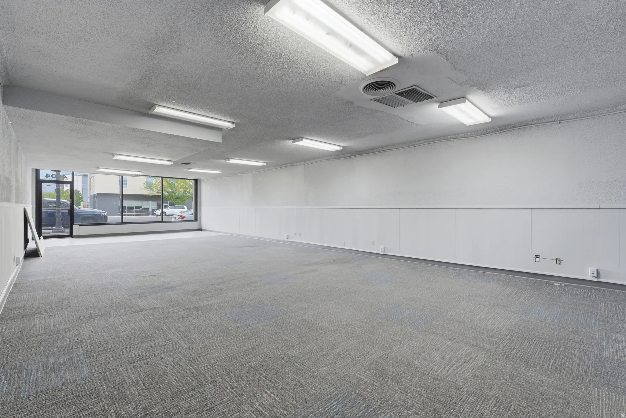 Expansive open-concept commercial space featuring large storefront windows, textured gray carpeting, white walls with wainscoting, and fluorescent overhead lighting